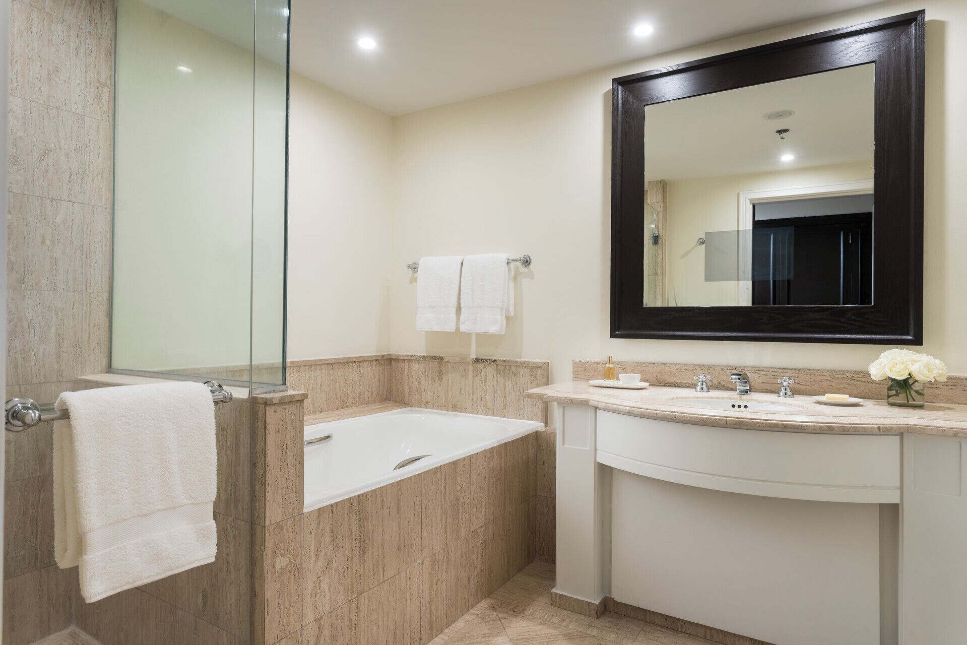 A bathroom with a glass shower enclosure, a bathtub, a sink with a mirror, and a vase with flowers.