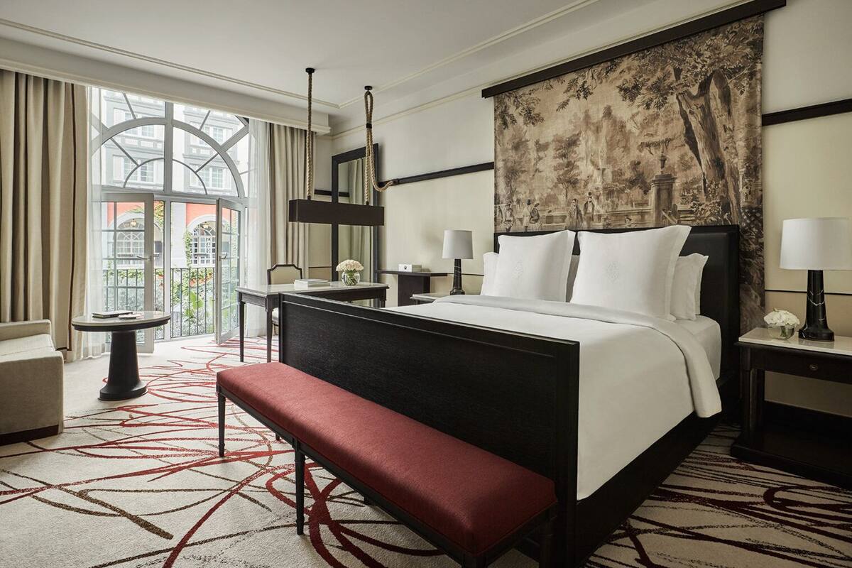 Habitación de hotel con una cama grande, un banco, un escritorio, una silla, una mesita de noche con lámpara y un ventanal con cortinas.