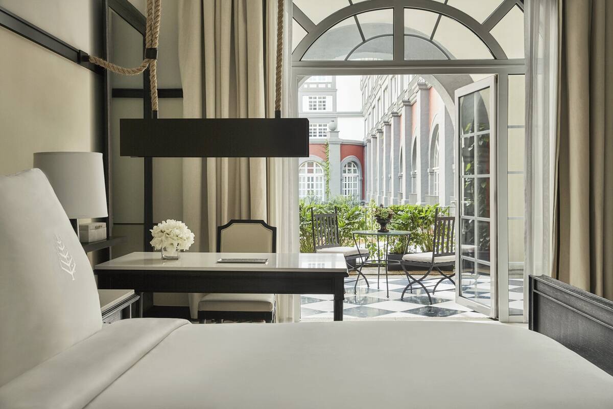 Habitación de hotel con cama, escritorio, silla, lámpara y vista a un patio con plantas y un edificio.