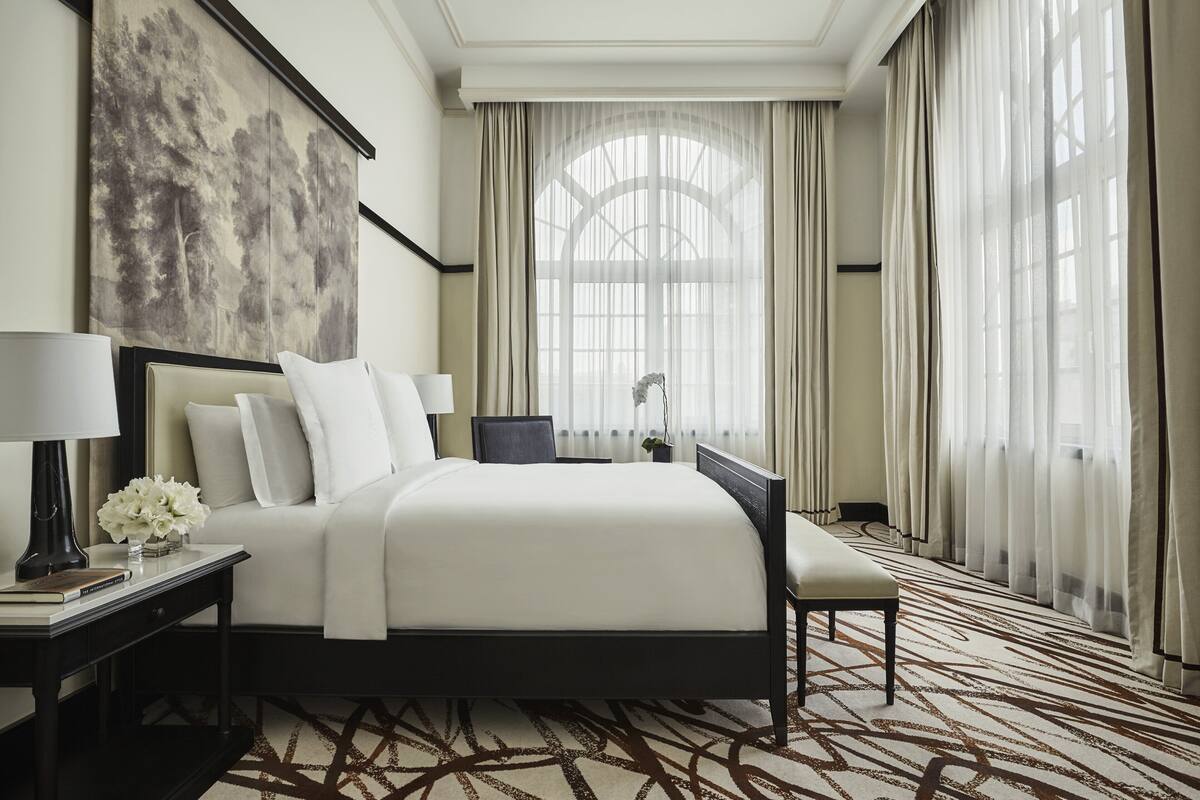 Habitación de hotel con una cama grande, una mesita de noche con lámpara, una silla y un ventanal con cortinas traslúcidas.