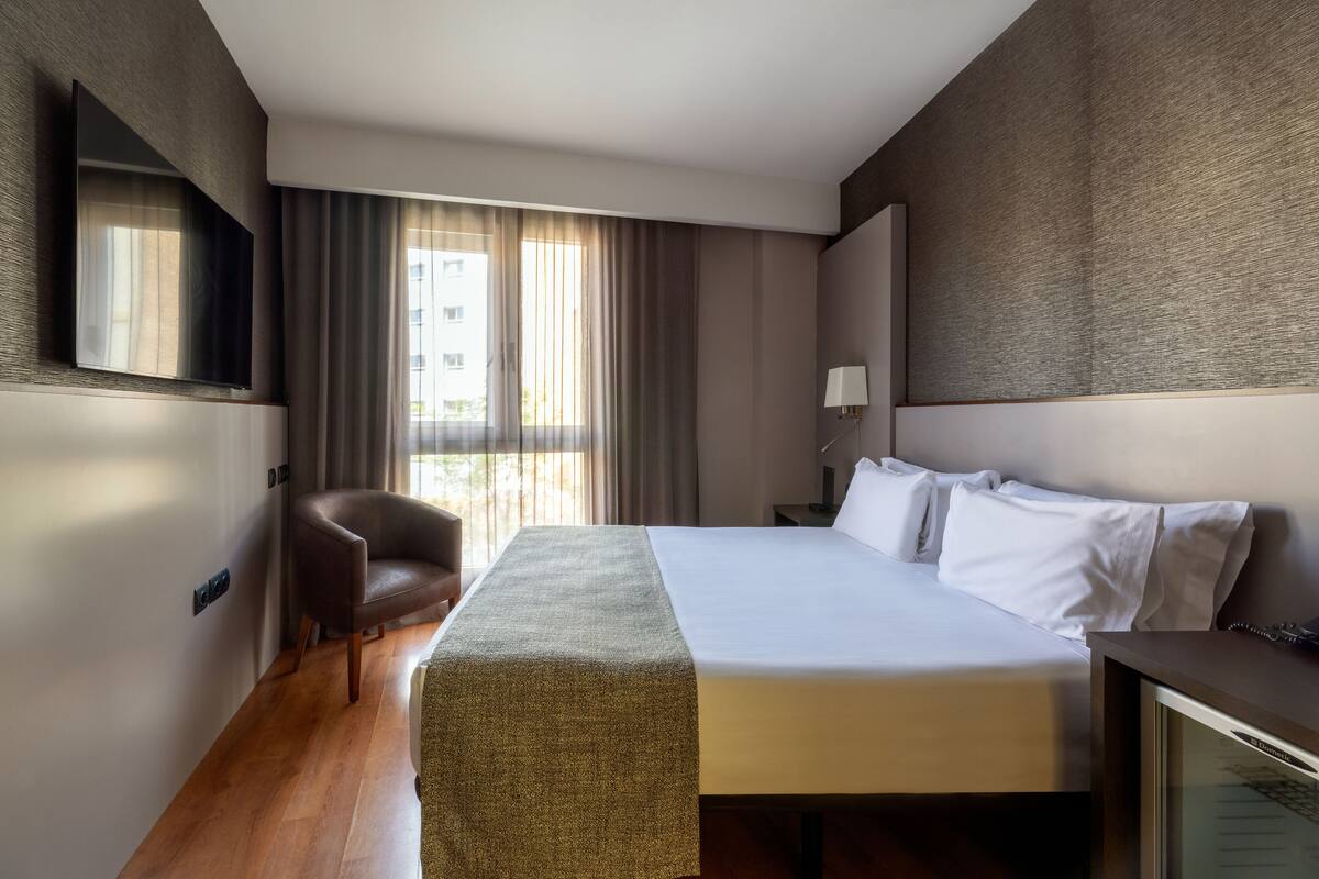 Ein modernes Hotelzimmer mit einem großen Bett, einem Schreibtisch, einem Sessel und einem an der Wand befestigten Fernseher.