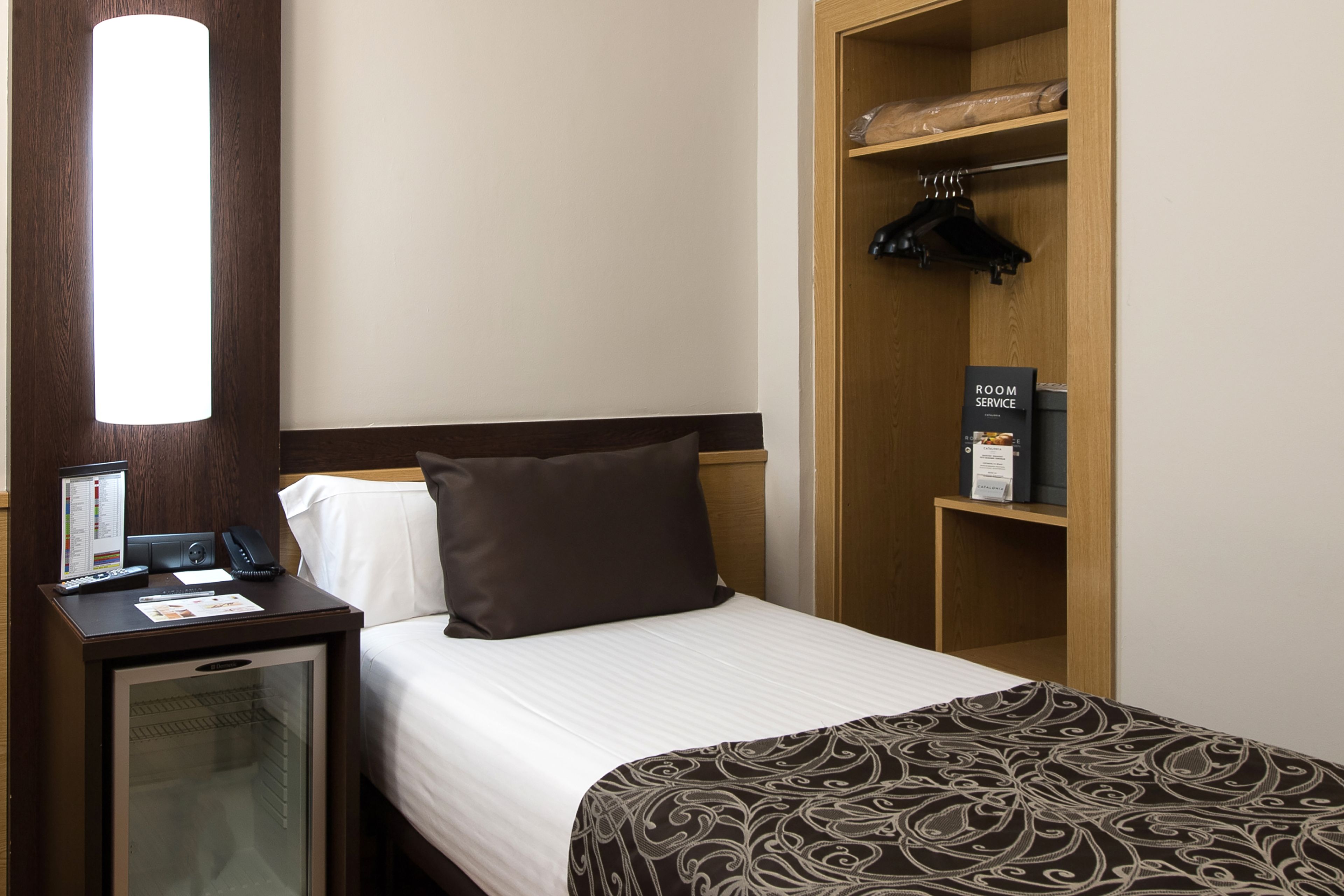 Habitación de hotel con cama, mesita de noche con teléfono y pequeño refrigerador, armario de madera con perchas y un letrero que indica servicio a la habitación.