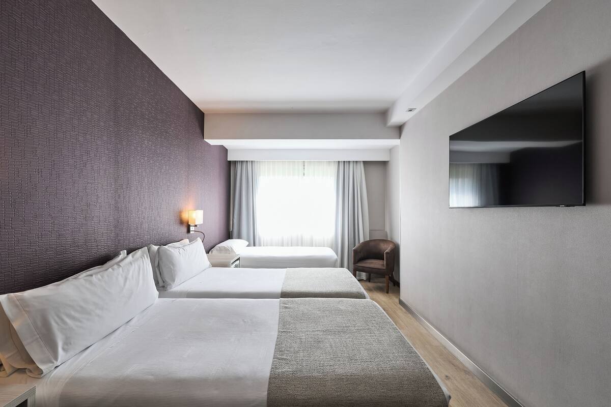 Ein Hotelzimmer mit zwei Betten, einem Flachbildfernseher, einem Sessel und einem Fenster mit Vorhängen.
