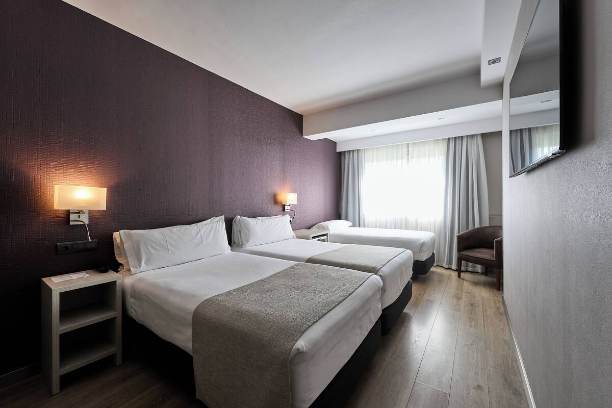 Ein Hotelzimmer mit zwei Betten, einem Nachttisch, einem Sessel, einem Fernseher und einem Fenster mit Vorhängen.