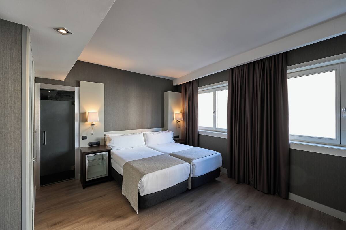Ein modernes Hotelzimmer mit Doppelbett, Nachttischen, einem Kleiderschrank und einem großen Fenster mit Vorhängen.