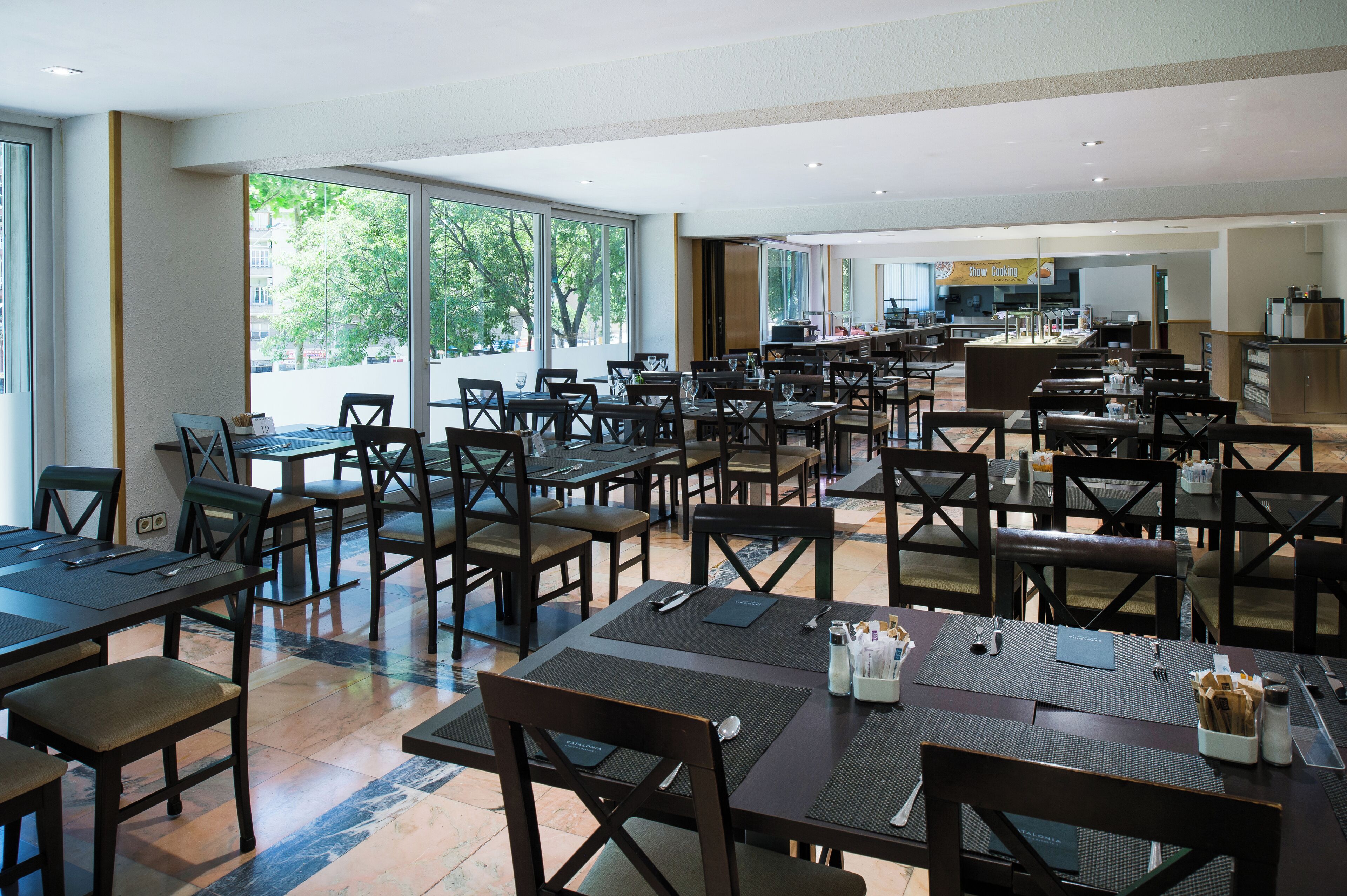 A spacious dining area with multiple tables and chairs, large windows providing natural light, and a kitchen area in the background.