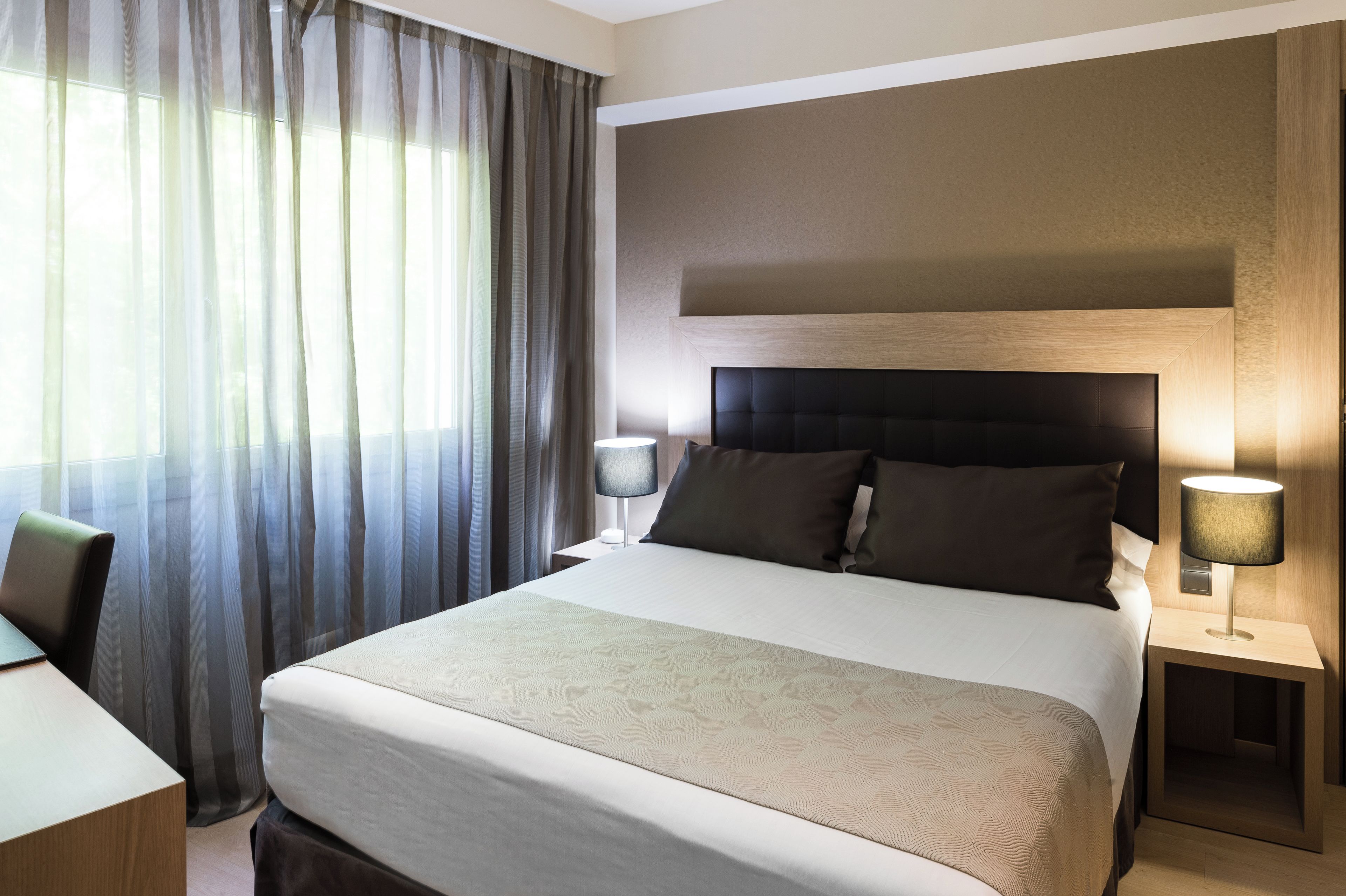 A hotel room with a large bed, two bedside lamps, a chair, and a window with curtains.
