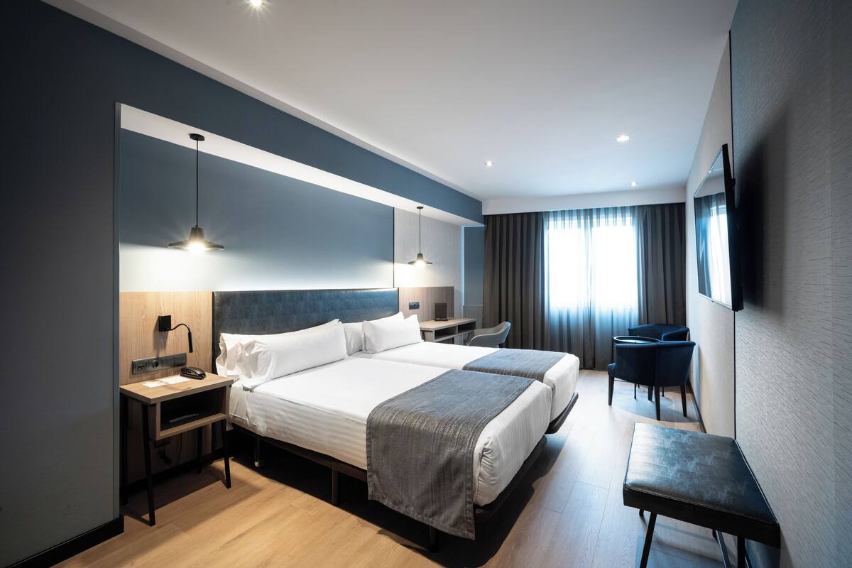 Ein modernes Hotelzimmer mit zwei Betten, einem Schreibtisch, einem Stuhl und einem an der Wand befestigten Fernseher.