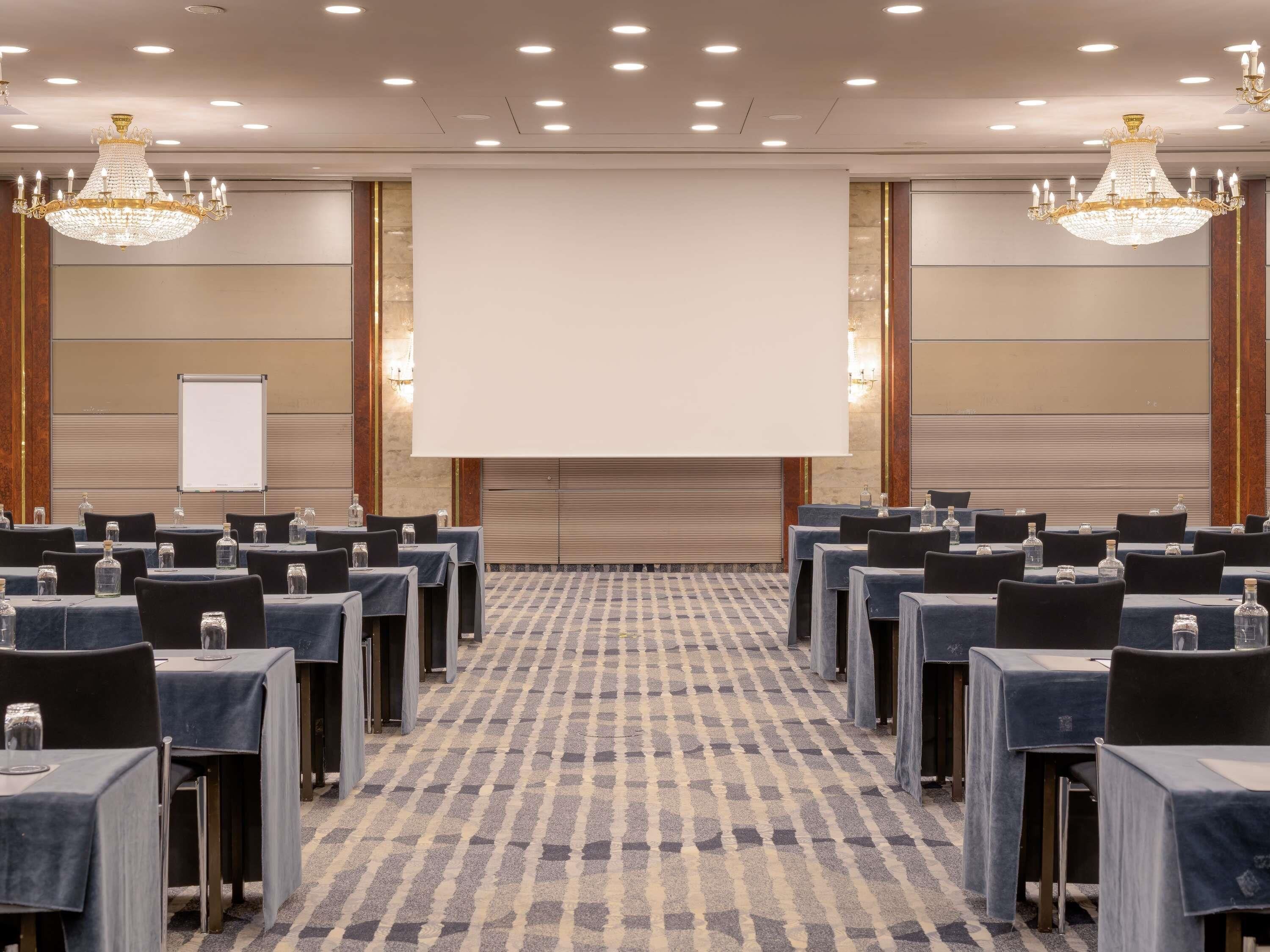 Een congreszaal met rijen tafels, een projectiescherm en kroonluchters.