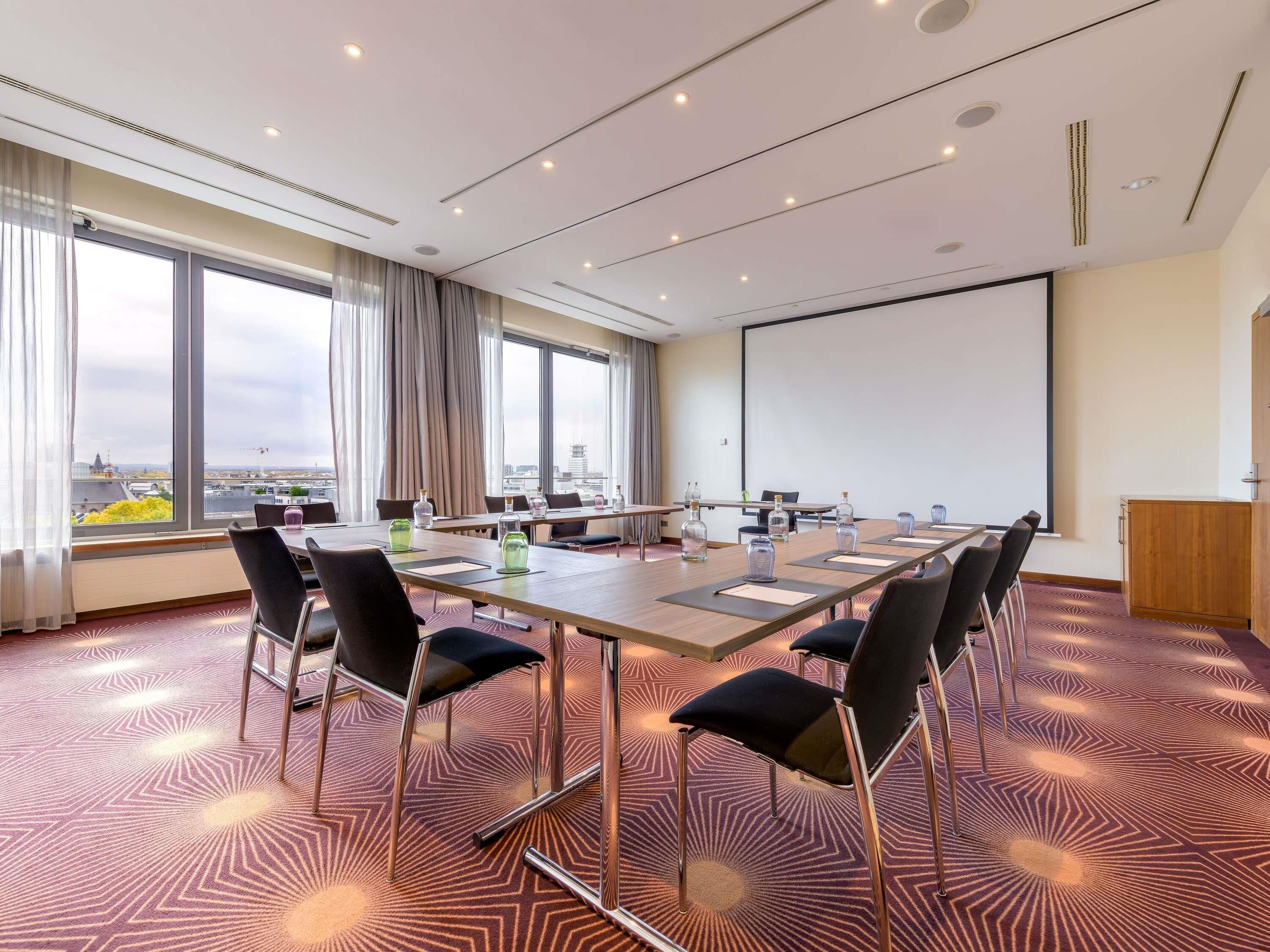 Une salle de conférence équipée d’une grande table, de chaises, d’un écran de projection et de grandes fenêtres avec des rideaux.