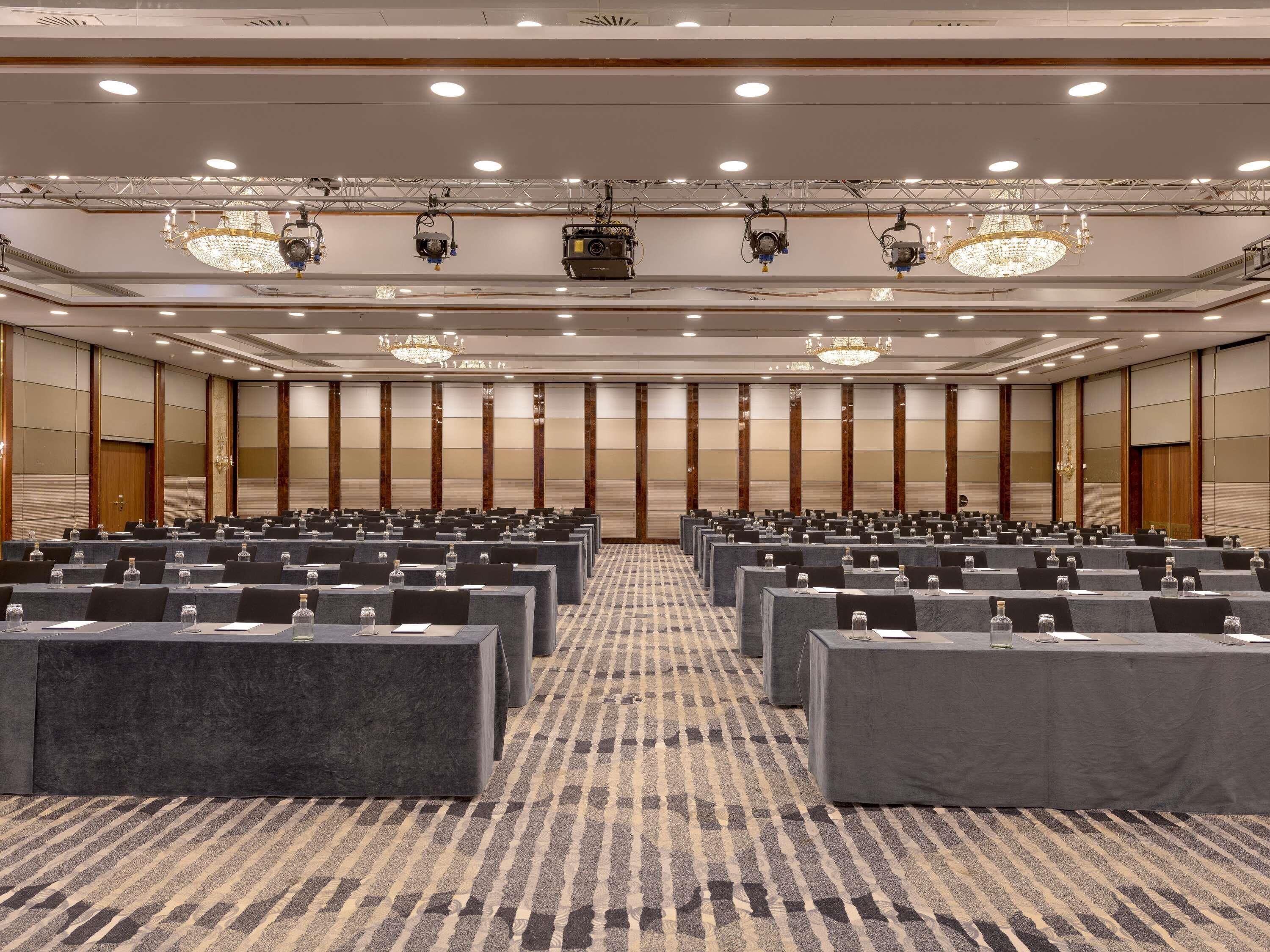 A conference room with rows of tables, overhead lighting, and a patterned carpet.