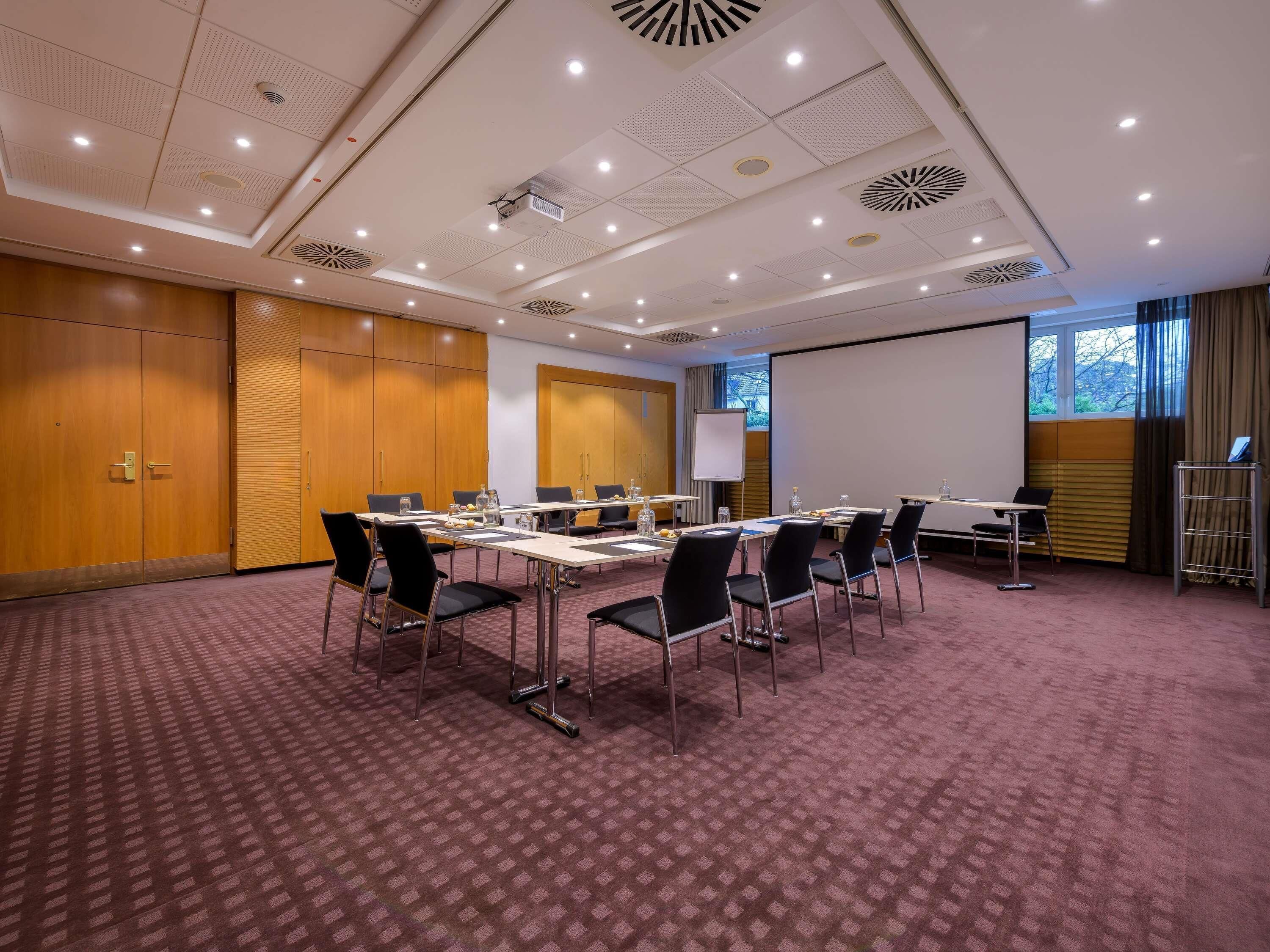 Ein Konferenzraum mit großem Tisch, Stühlen, einer Projektionsleinwand und einem Präsentationsbildschirm.