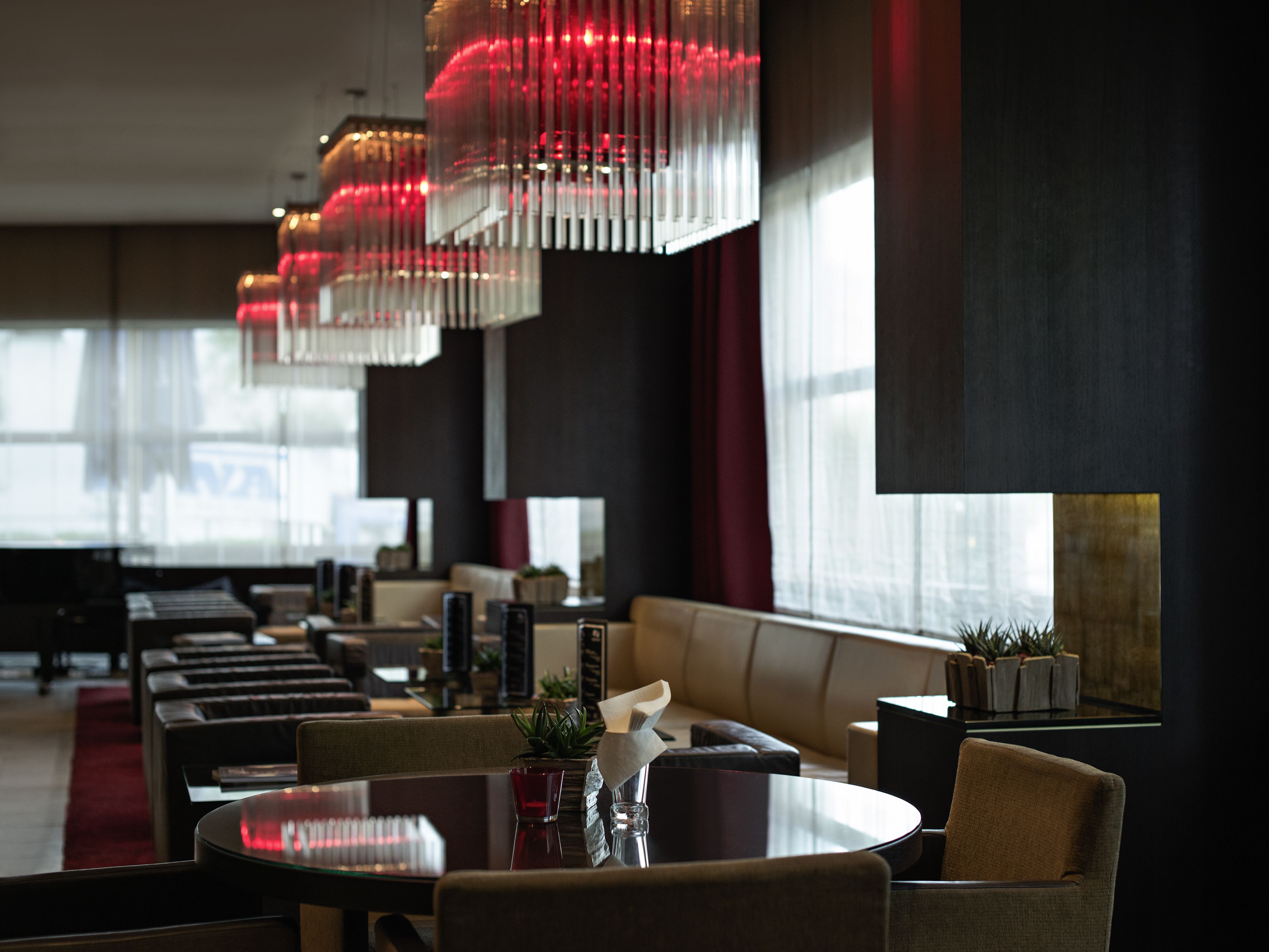 A modern dining area with round tables and cushioned seating, large windows with curtains, and decorative red lighting.