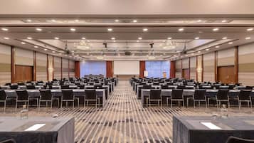 Une salle de conférence avec des rangées de tables, des chaises et une scène équipée d’un écran de projection.