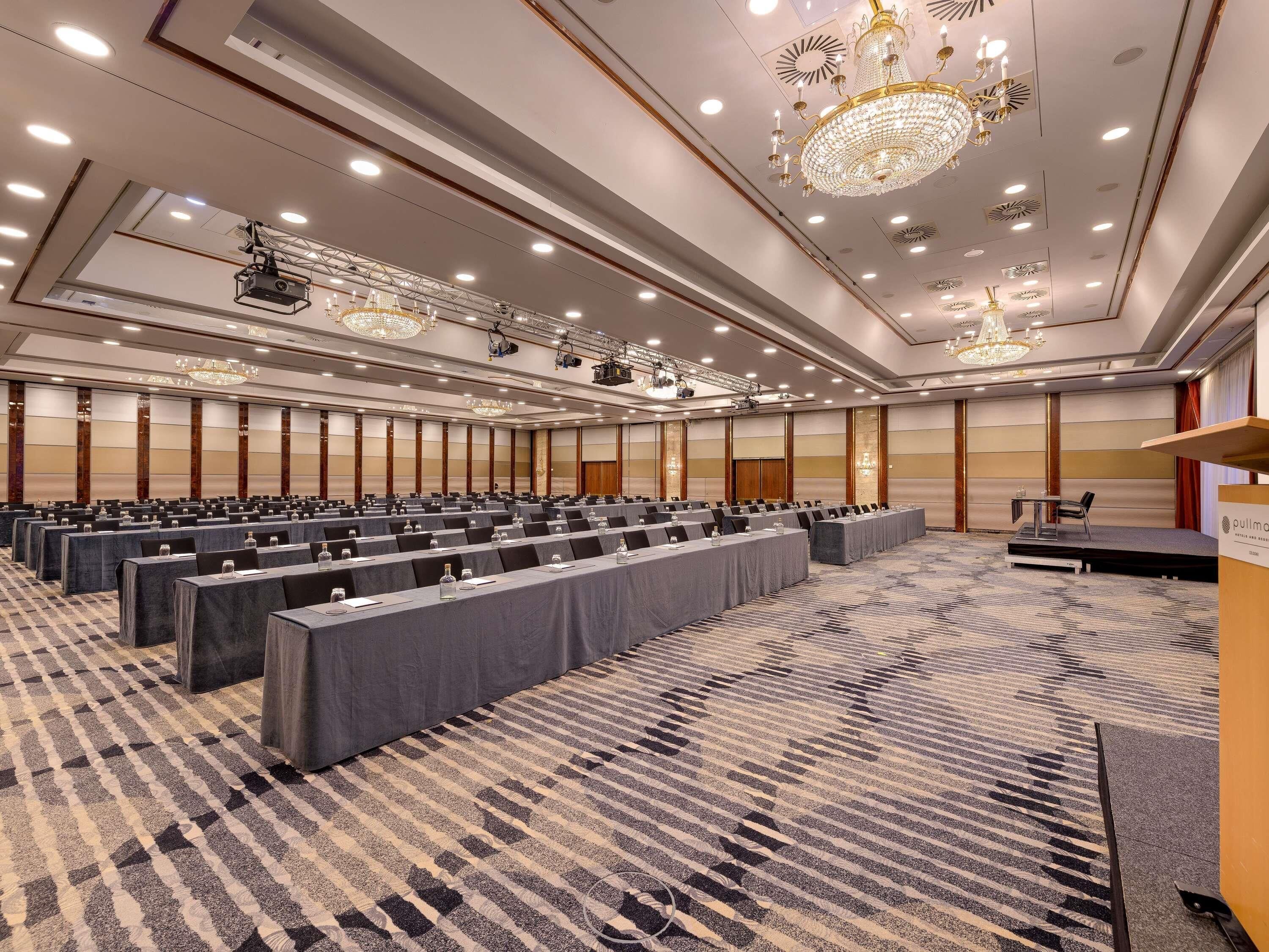 A large conference room with rows of tables, a stage with a podium, and chandeliers.