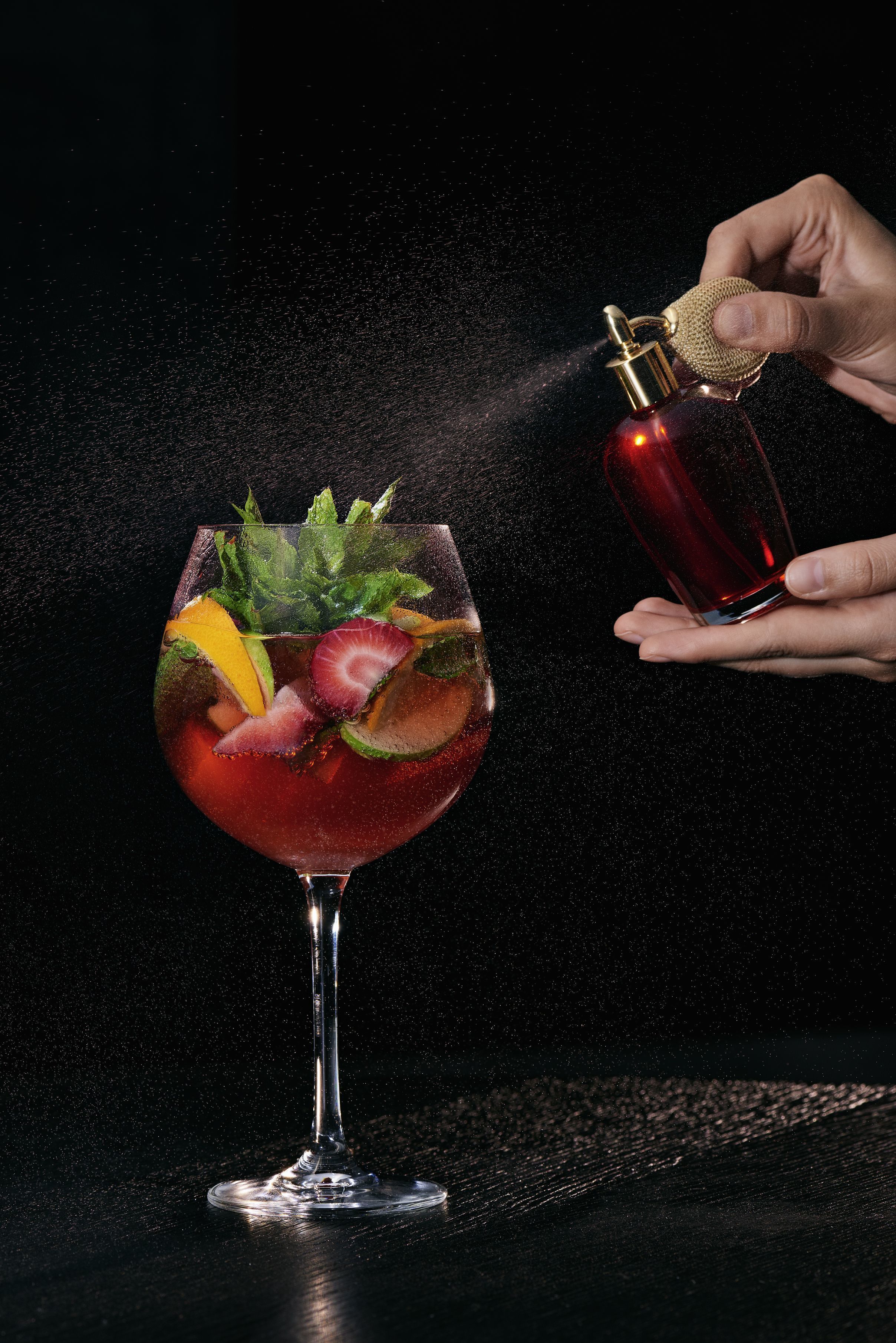 Une main tenant une bouteille contenant un liquide rouge, et en vaporisant une partie sur un verre contenant un cocktail aux fruits et à la menthe.