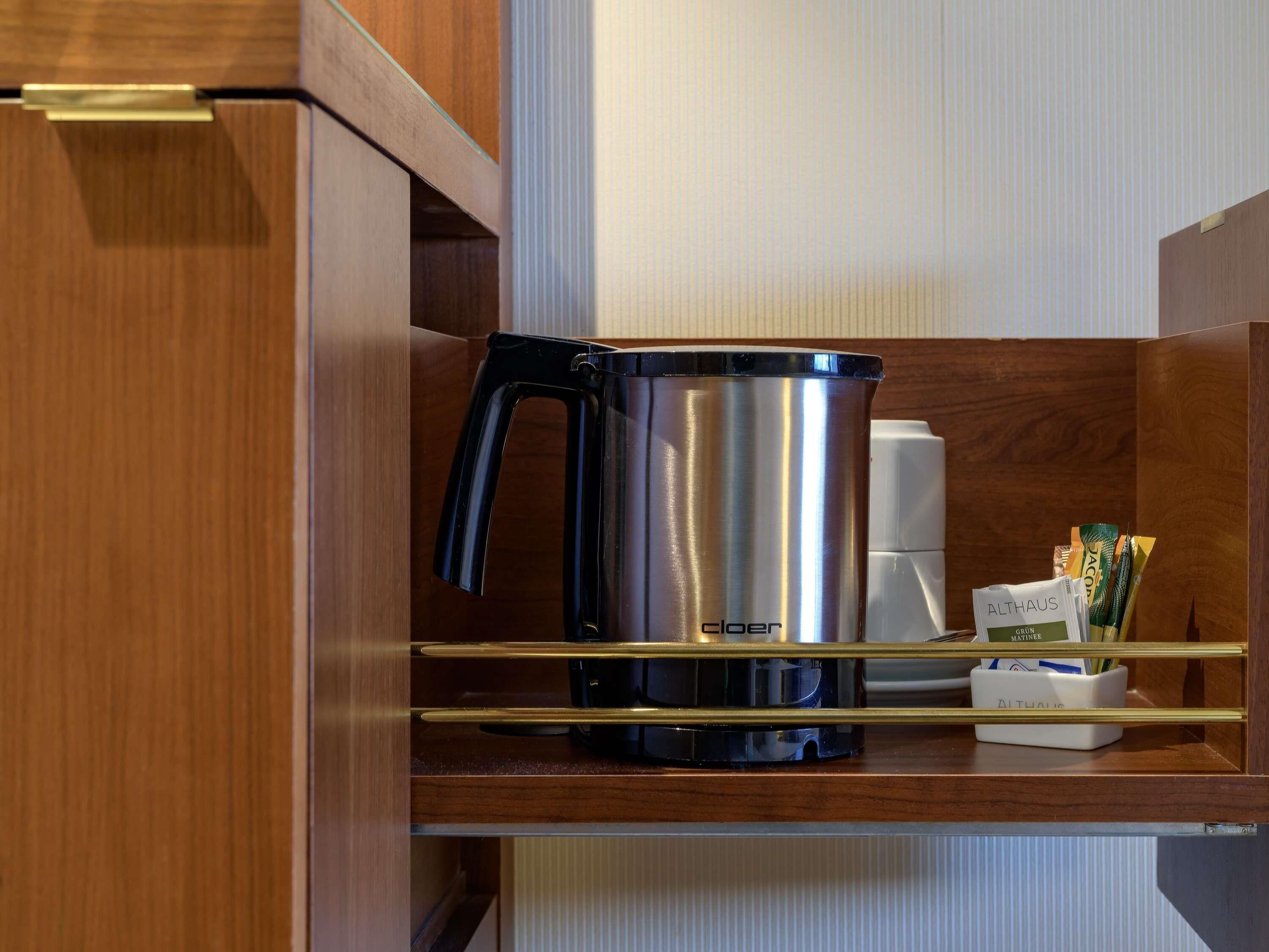 A stainless steel coffee maker on a wooden shelf with a white coffee maker and a container holding pens and pencils.