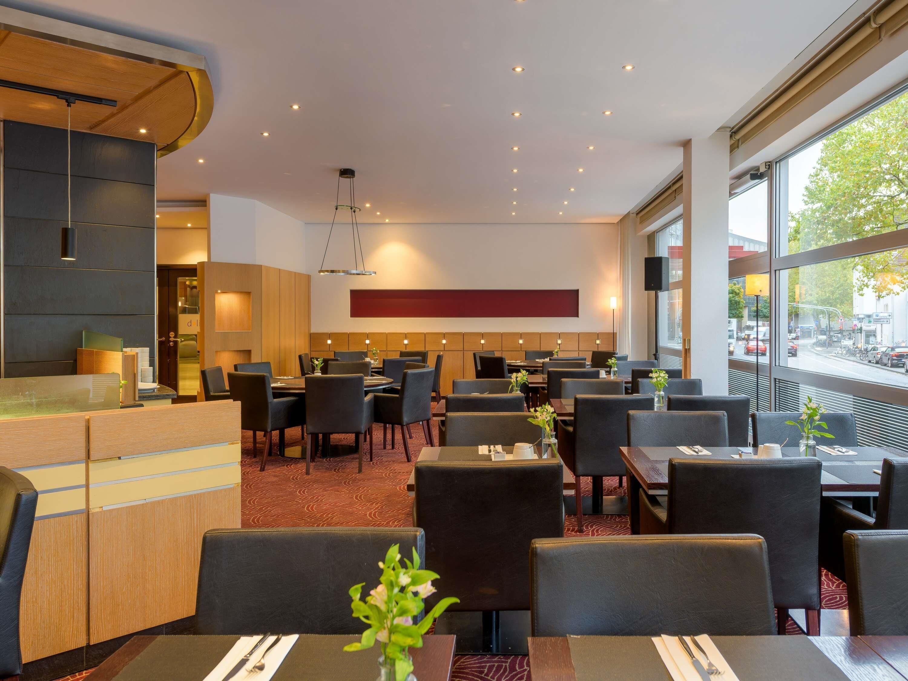 Un espace de restauration moderne dans un hôtel, avec de nombreuses tables et chaises, un comptoir d’accueil et de grandes fenêtres donnant sur la rue.
