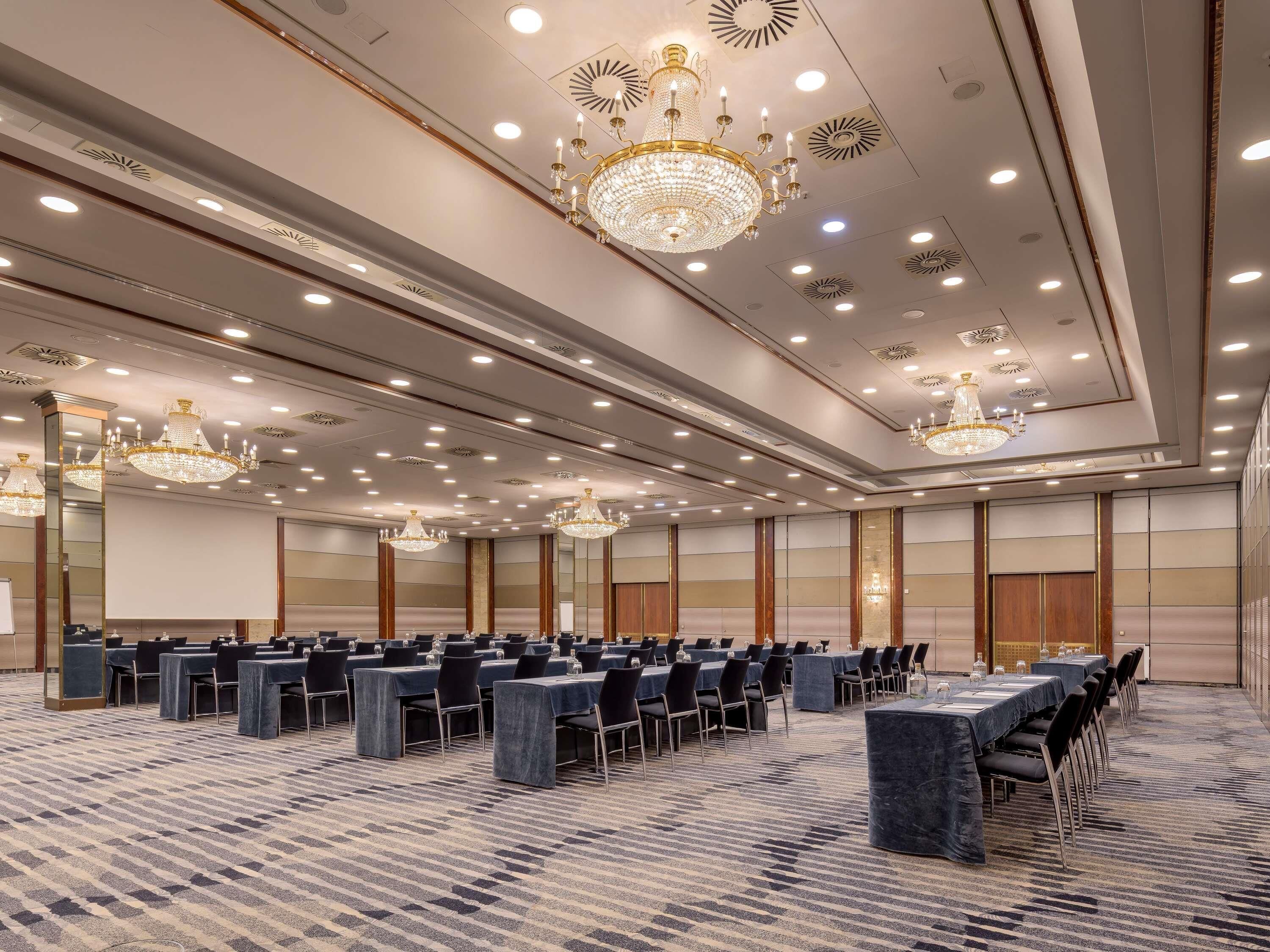 Une grande salle de conférence avec de longues rangées de tables et de chaises, des lustres et des spots encastrés dans le plafond.
