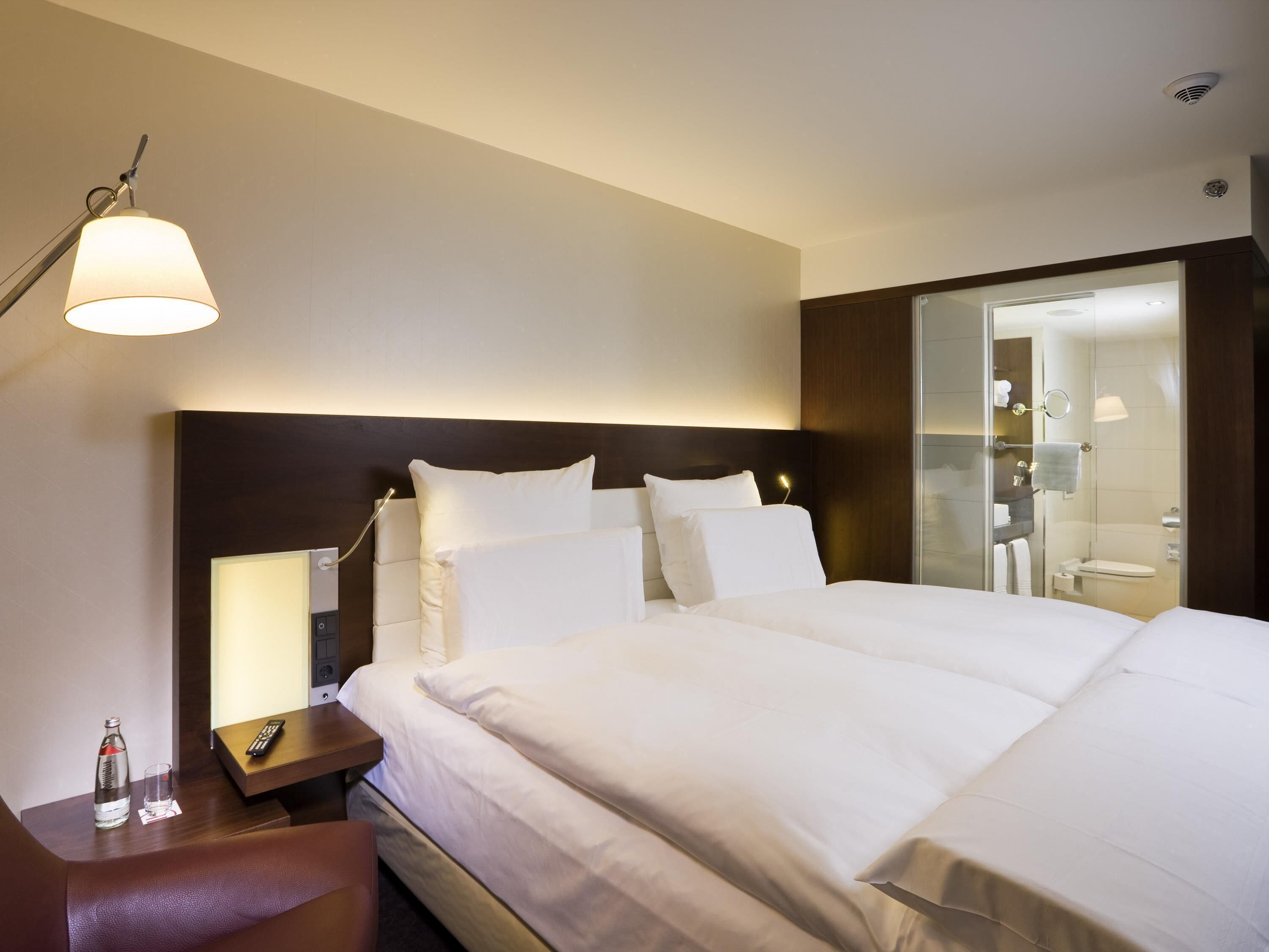 A hotel room with a bed, bedside table, lamp, and a view of the bathroom.