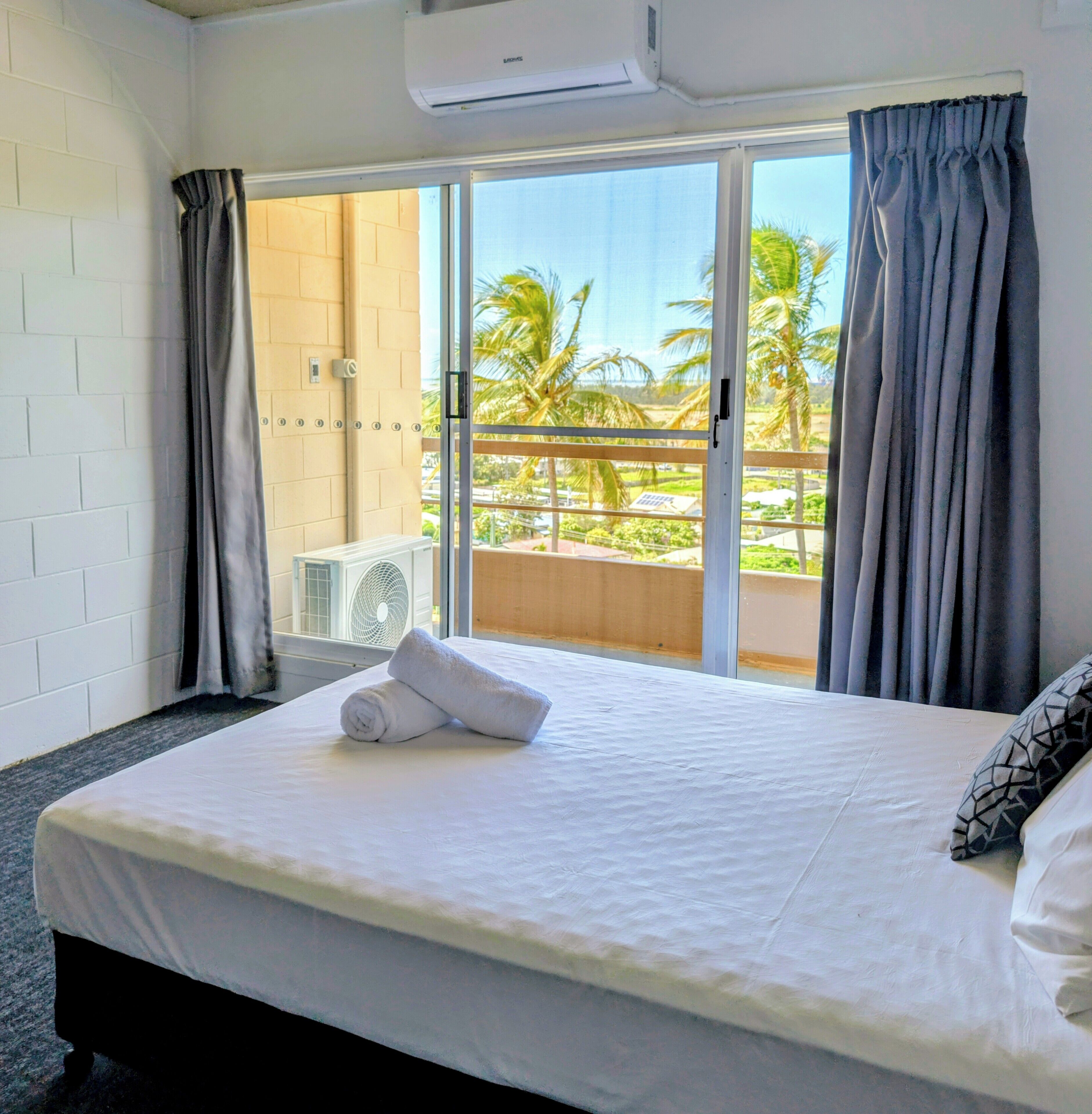 Une chambre d’hôtel avec un lit, une fenêtre avec des rideaux, un appareil de climatisation et une vue sur des palmiers.
