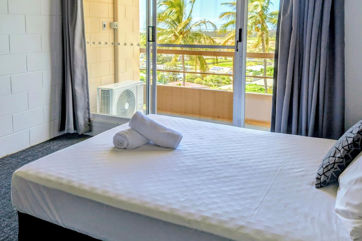 A hotel room with a bed, a window with curtains, an air conditioning unit, and a view of palm trees.