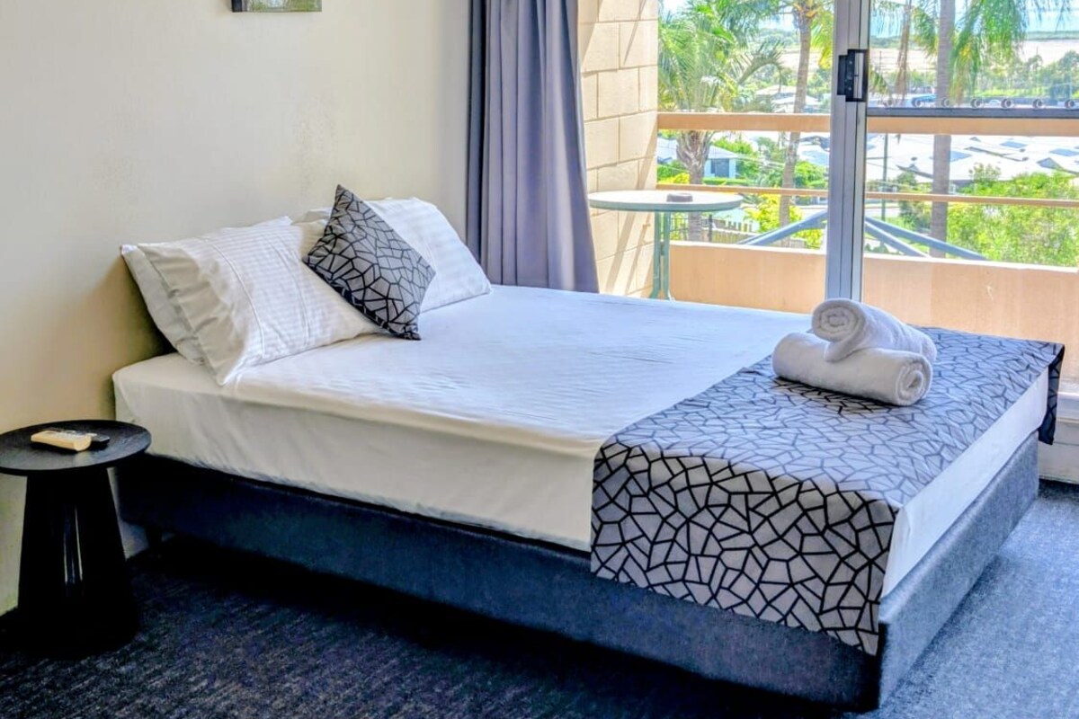 Une chambre d’hôtel équipée d’un lit, d’une table de chevet, d’un ventilateur de plafond, d’un climatiseur, d’une fenêtre avec des rideaux et offrant une vue sur des palmiers.