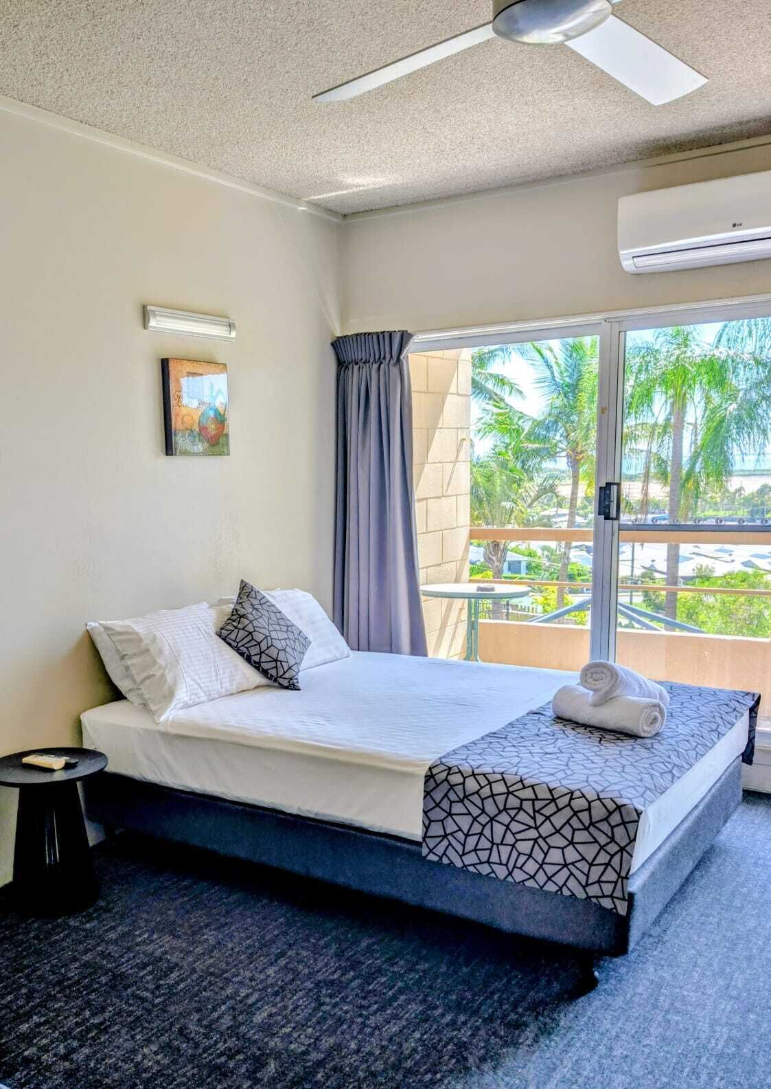 Une chambre d’hôtel équipée d’un lit, d’une table de chevet, d’un ventilateur de plafond, d’un climatiseur, d’une fenêtre avec des rideaux et offrant une vue sur des palmiers.