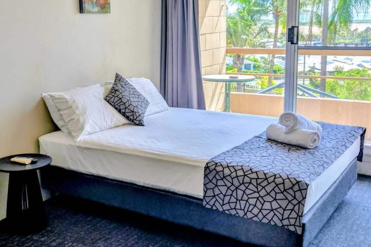A hotel room with a bed, a nightstand, a ceiling fan, an air conditioning unit, a window with curtains, and a view of palm trees.