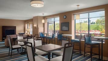 A hotel room with a TV, dining area with tables and chairs, large windows with a view of trees and flowers, and a clock on the wall.
