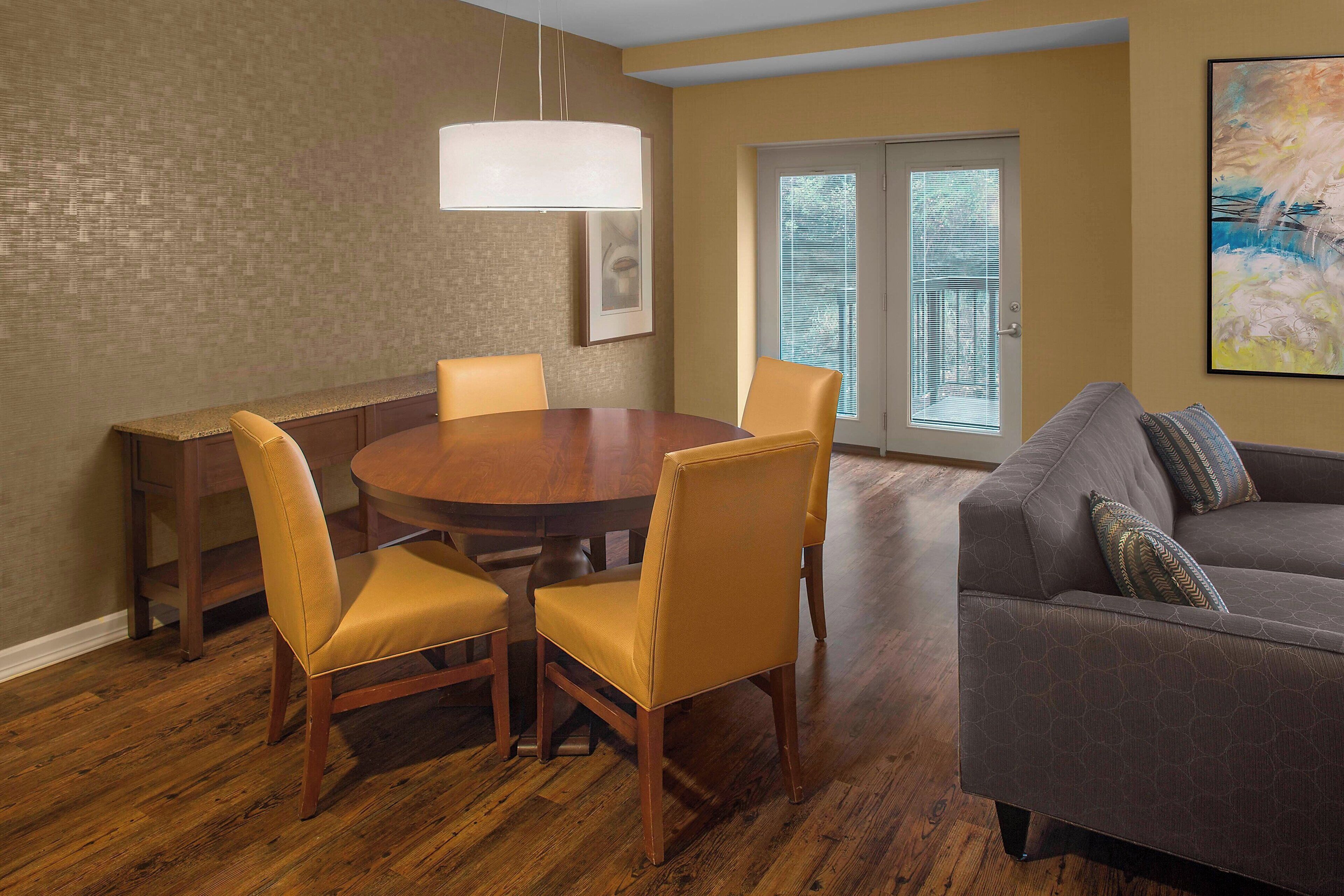 A dining area with a round table and four chairs, a sofa, and a large window with blinds.