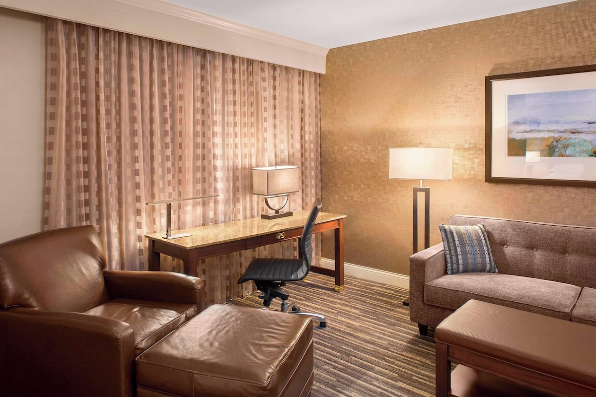Une chambre d’hôtel avec un canapé en cuir marron, un fauteuil en cuir marron, un bureau en bois avec une chaise, un lampadaire et un tableau encadré au mur.