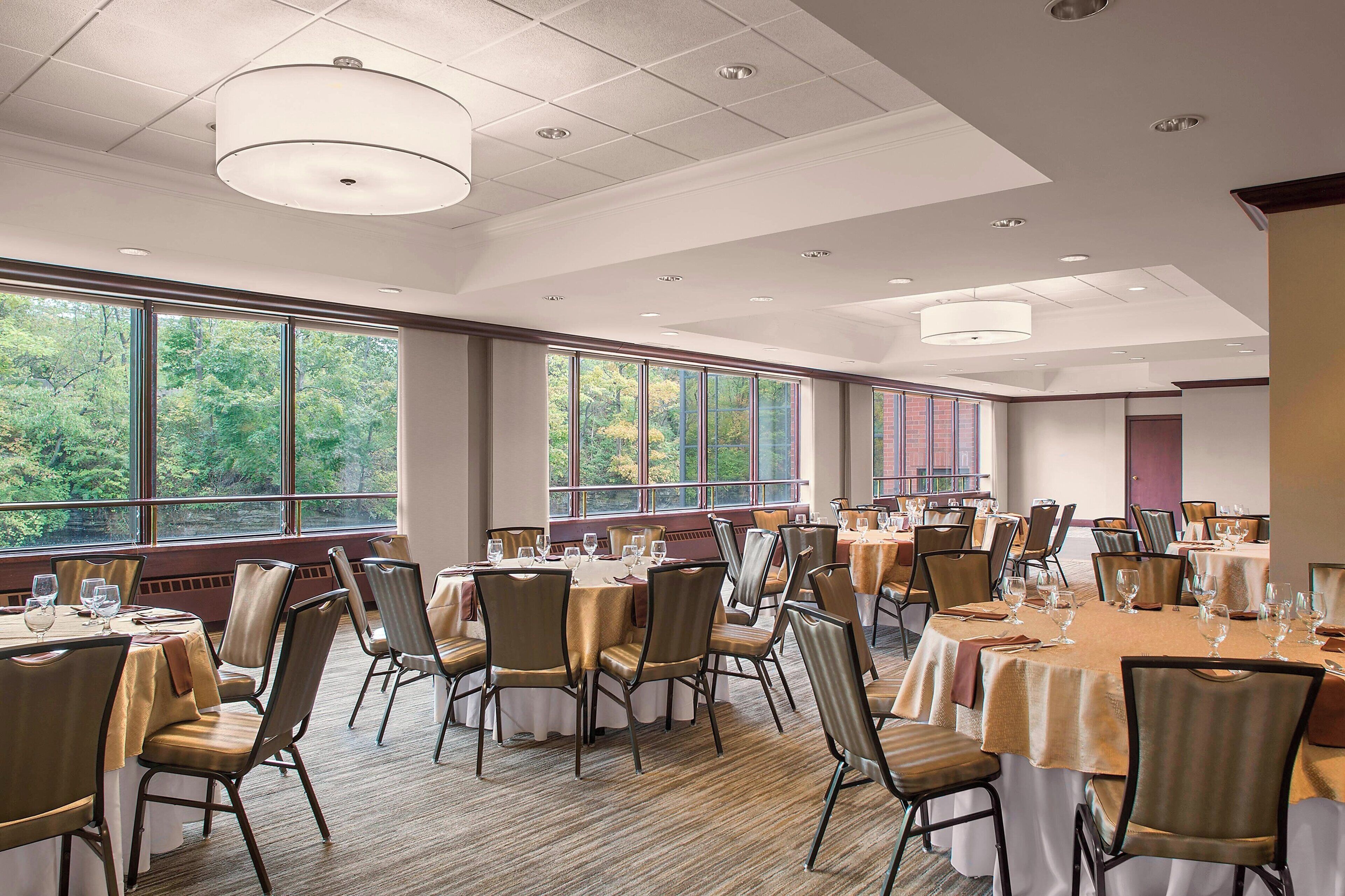Un vaste espace dédié à la salle à manger, avec de nombreuses tables et chaises, de grandes fenêtres offrant une vue sur les arbres et un design moderne au niveau du plafond.