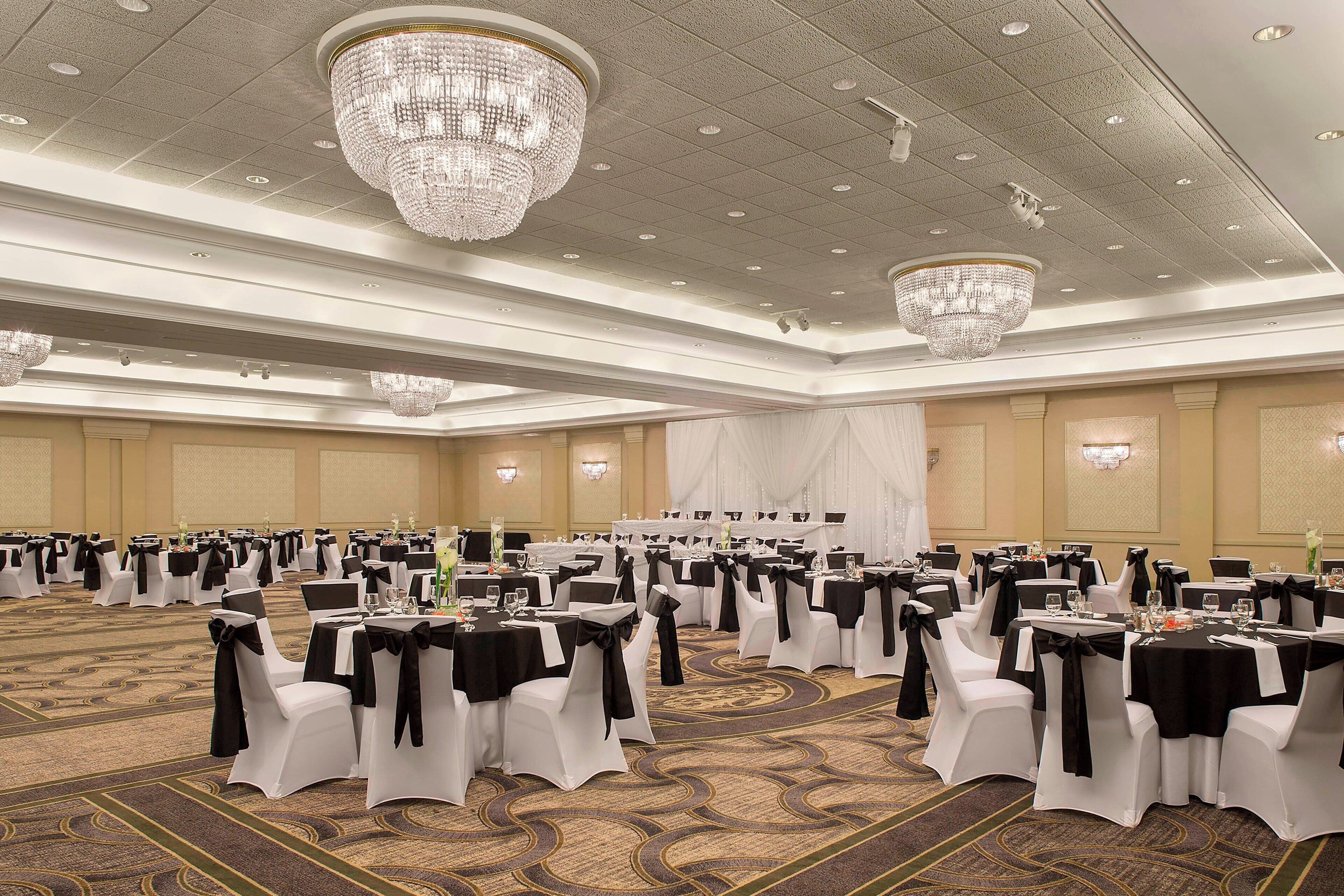 A banquet hall with round tables, white chairs, and black sashes, featuring chandeliers and a carpeted floor.