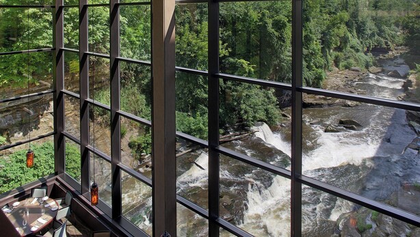 Restaurante con vista a un río y un bosque a través de amplios ventanales.