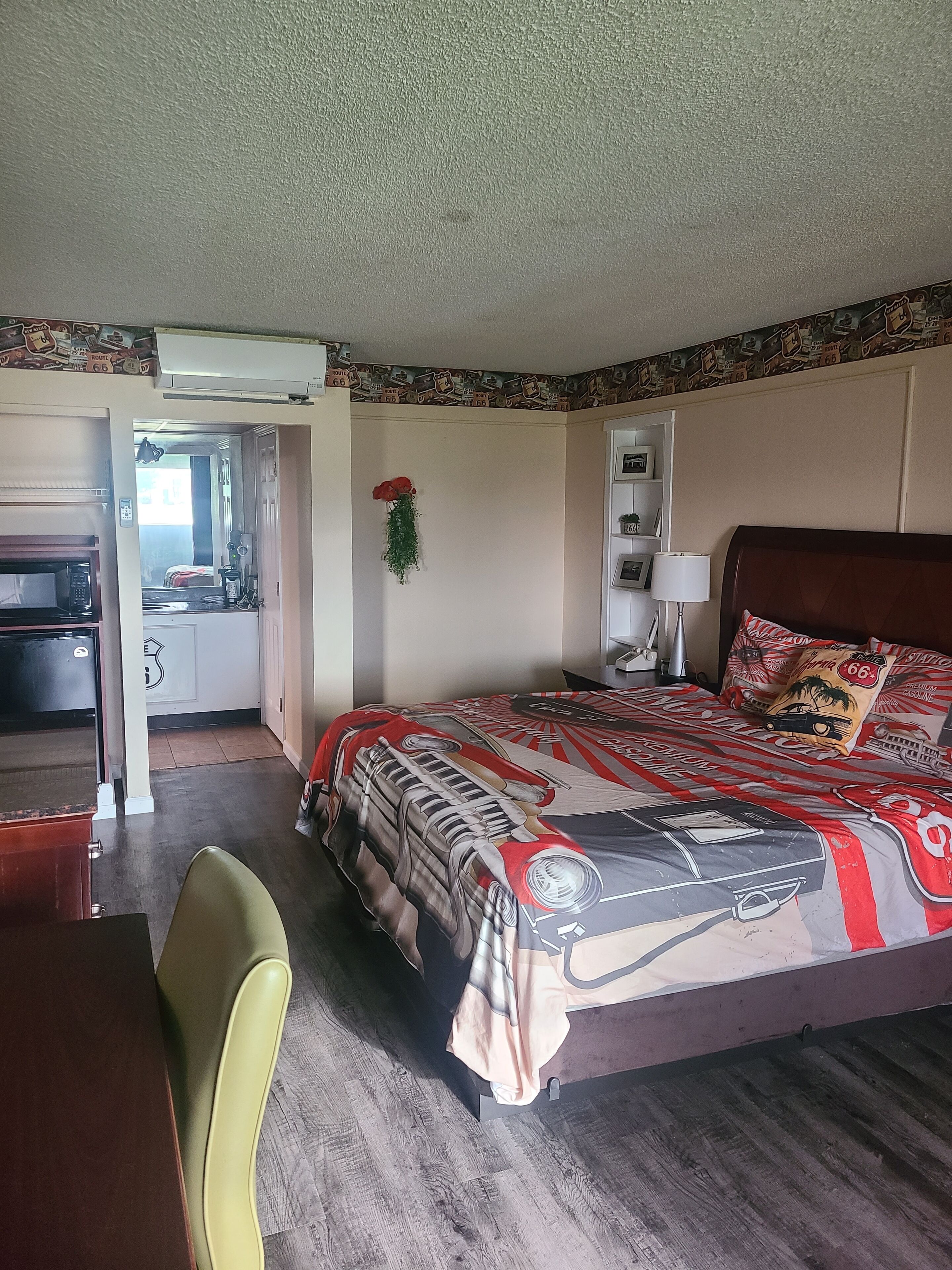 A hotel room with a bed, a desk, a chair, a microwave, a lamp, and a small shelf with decorative items.