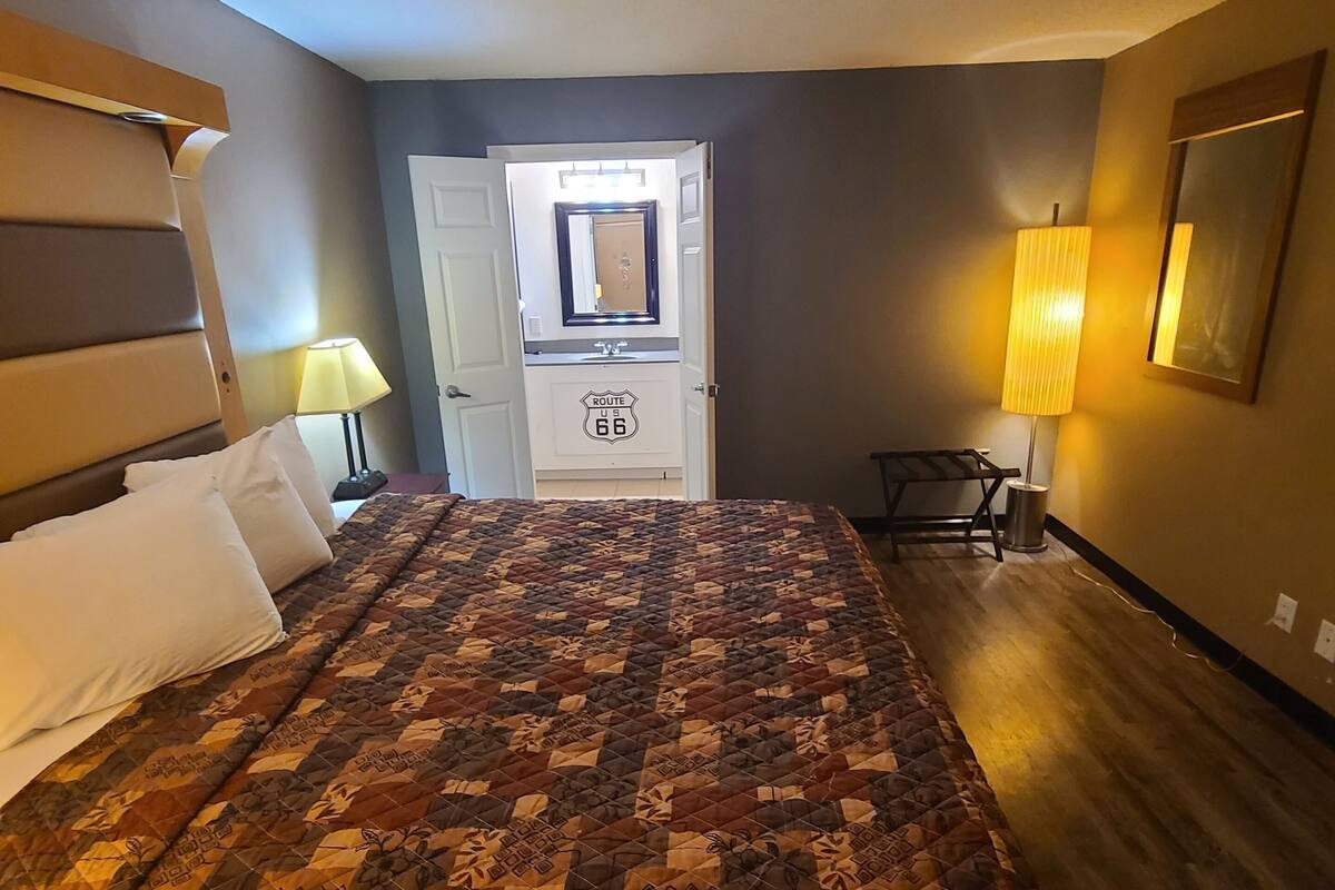 A hotel room with a bed, a desk, a lamp, a mirror, and a door leading to a hallway.