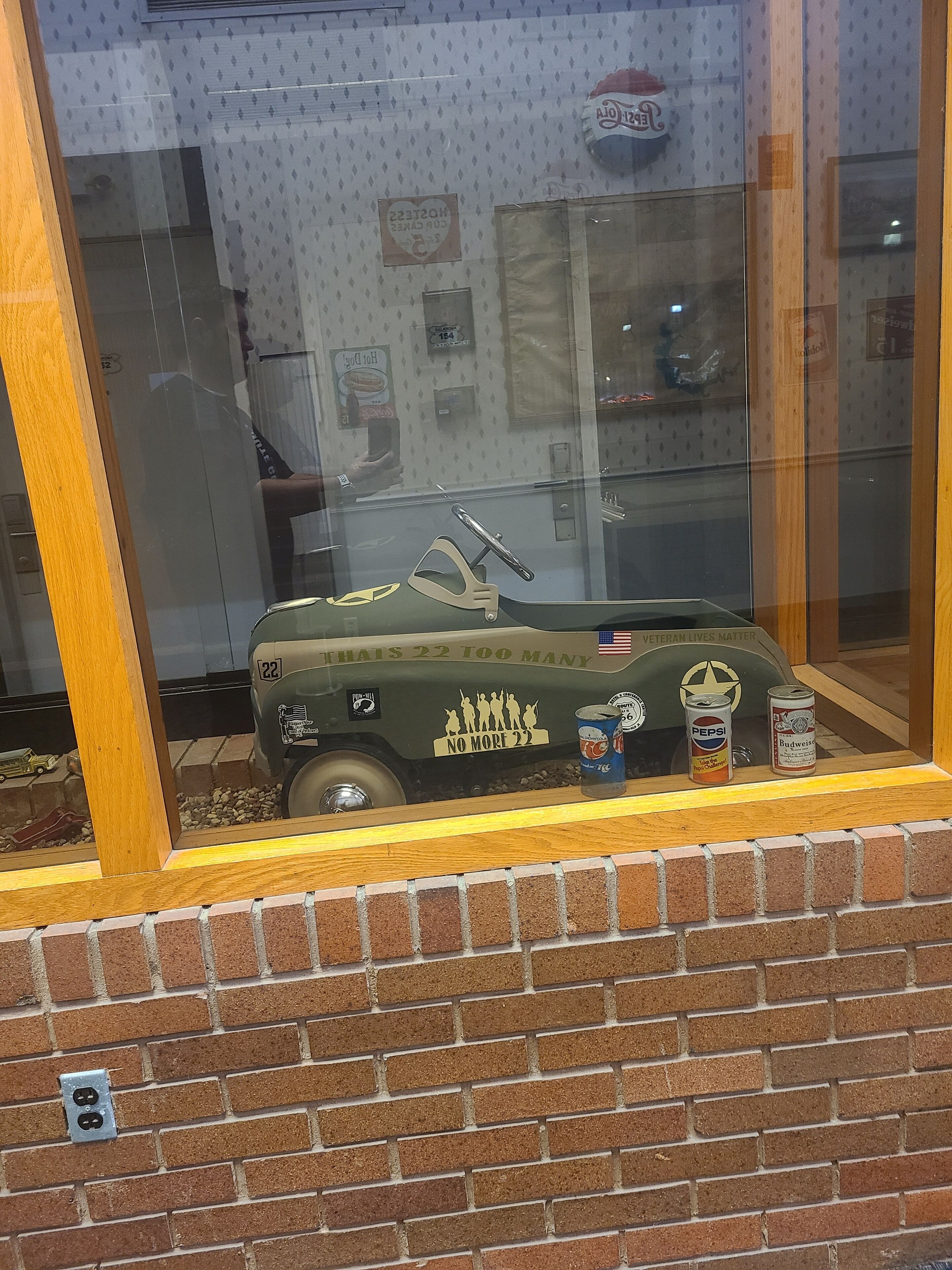 A model train set with a vintage car and two cans of soda on a brick wall.