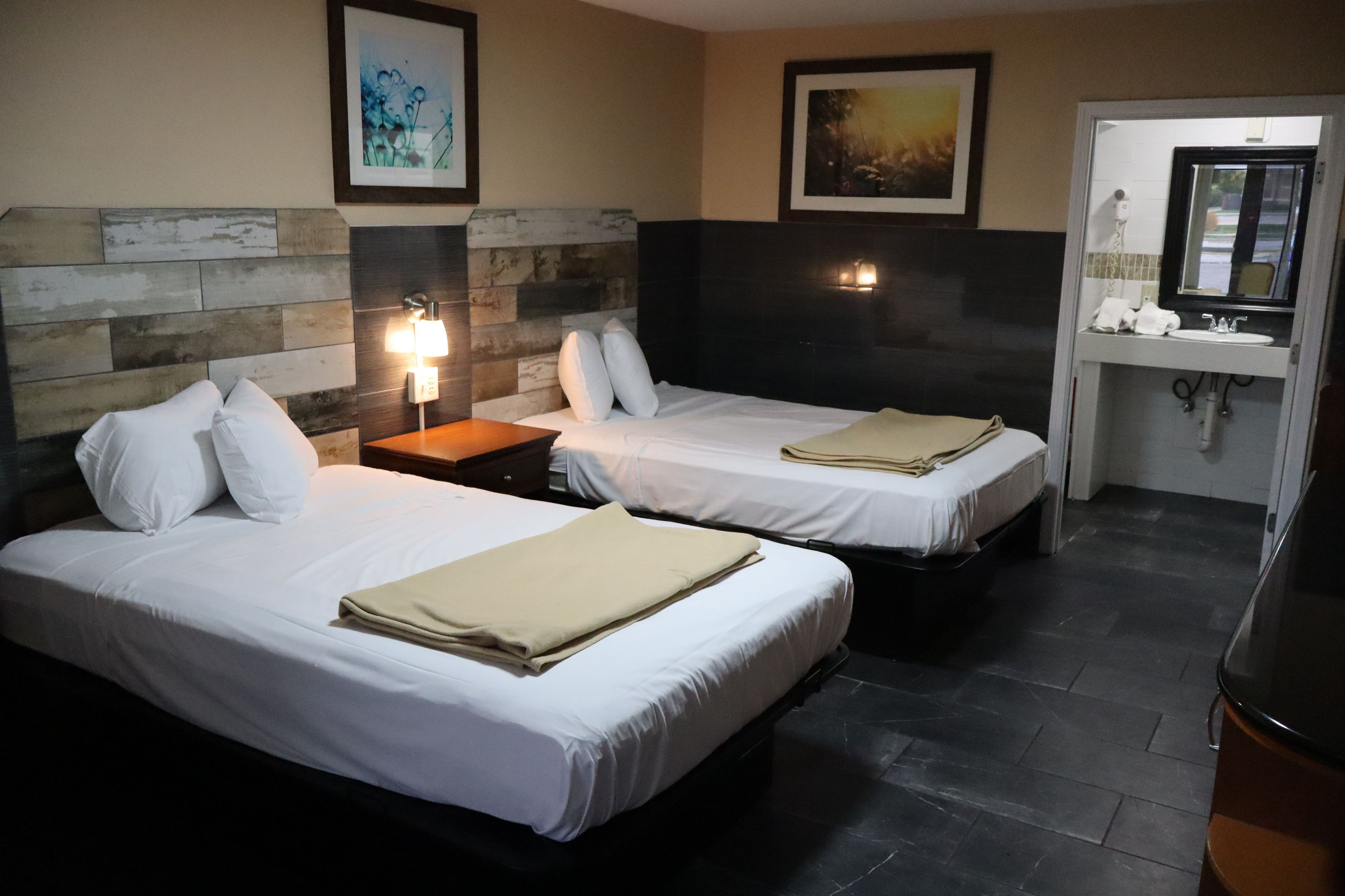 A hotel room with two beds, a nightstand, a wall-mounted lamp, and a bathroom with a sink and mirror.