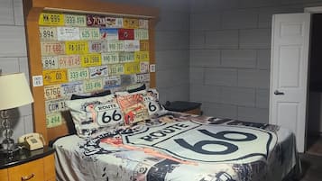 Un dormitorio con una cama cubierta con ropa de cama temática de la Ruta 66, un cabecero con varios números de matrícula y una mesita de noche con lámpara y teléfono.