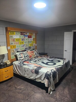 A bedroom with a bed covered in Route 66-themed bedding, a headboard with various license plates, and a nightstand with a lamp and phone.