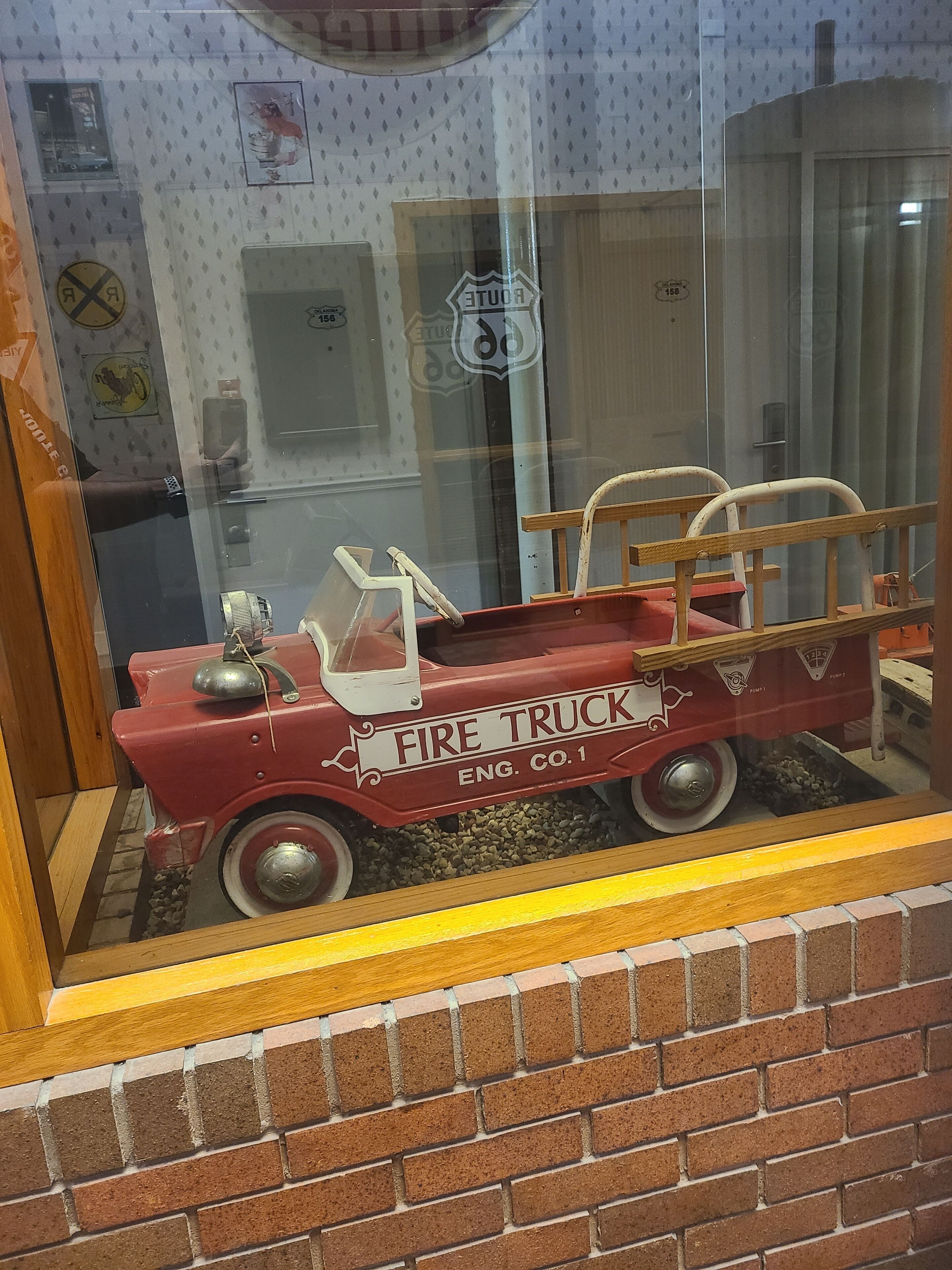 Une maquette de camion de pompiers exposée dans une vitrine, avec un mur en briques en arrière-plan.