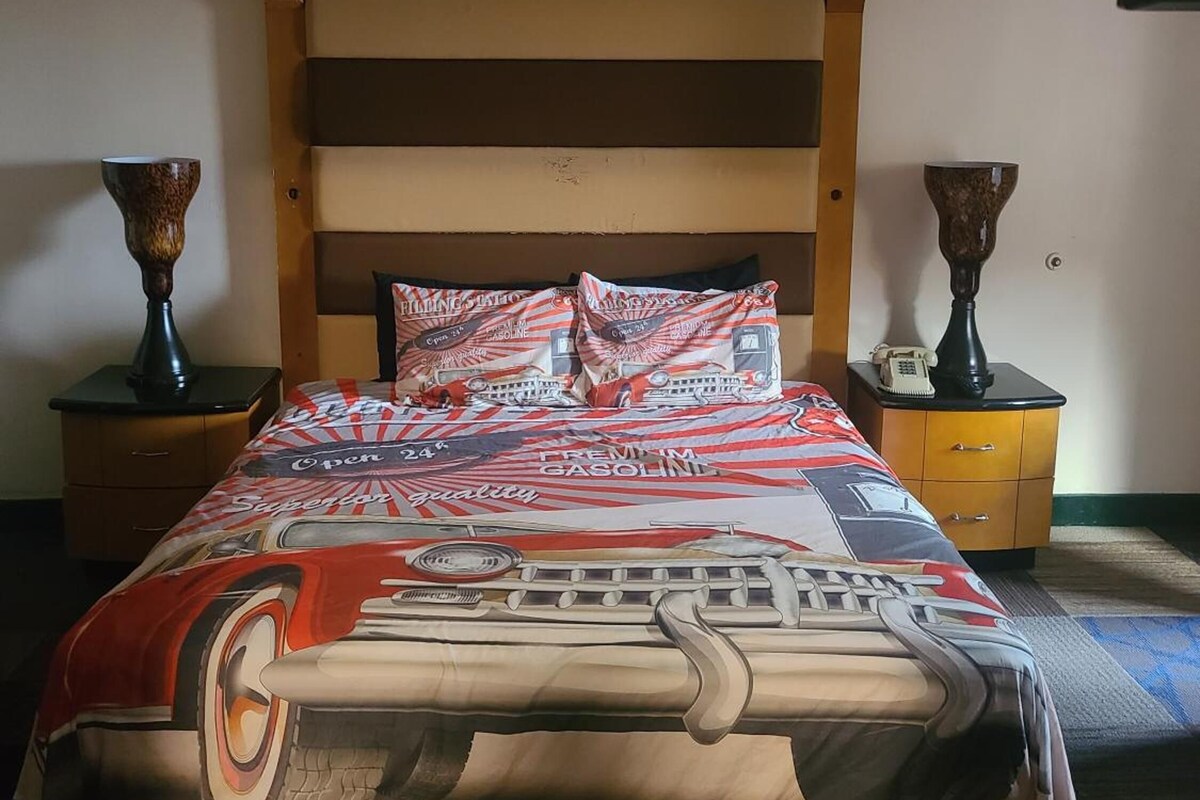Una cama con diseño de paisaje urbano, cabecera de madera, dos mesitas de noche con lámparas y suelo alfombrado.