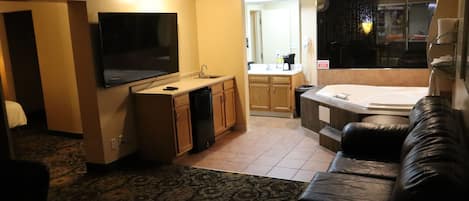 A hotel room with a flat-screen TV, kitchenette, a sofa, and a jacuzzi.