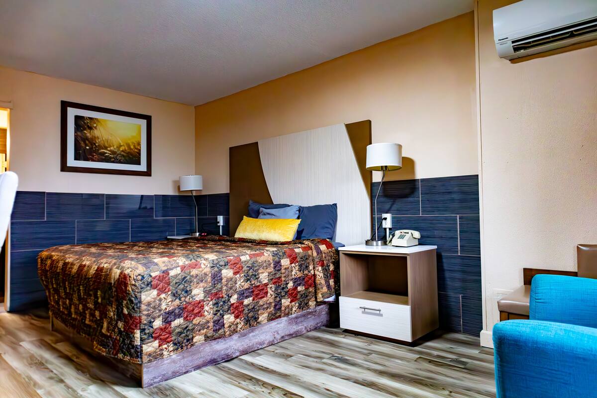A hotel room with a large bed, a nightstand with a lamp, a chair, and a wall-mounted air conditioner.
