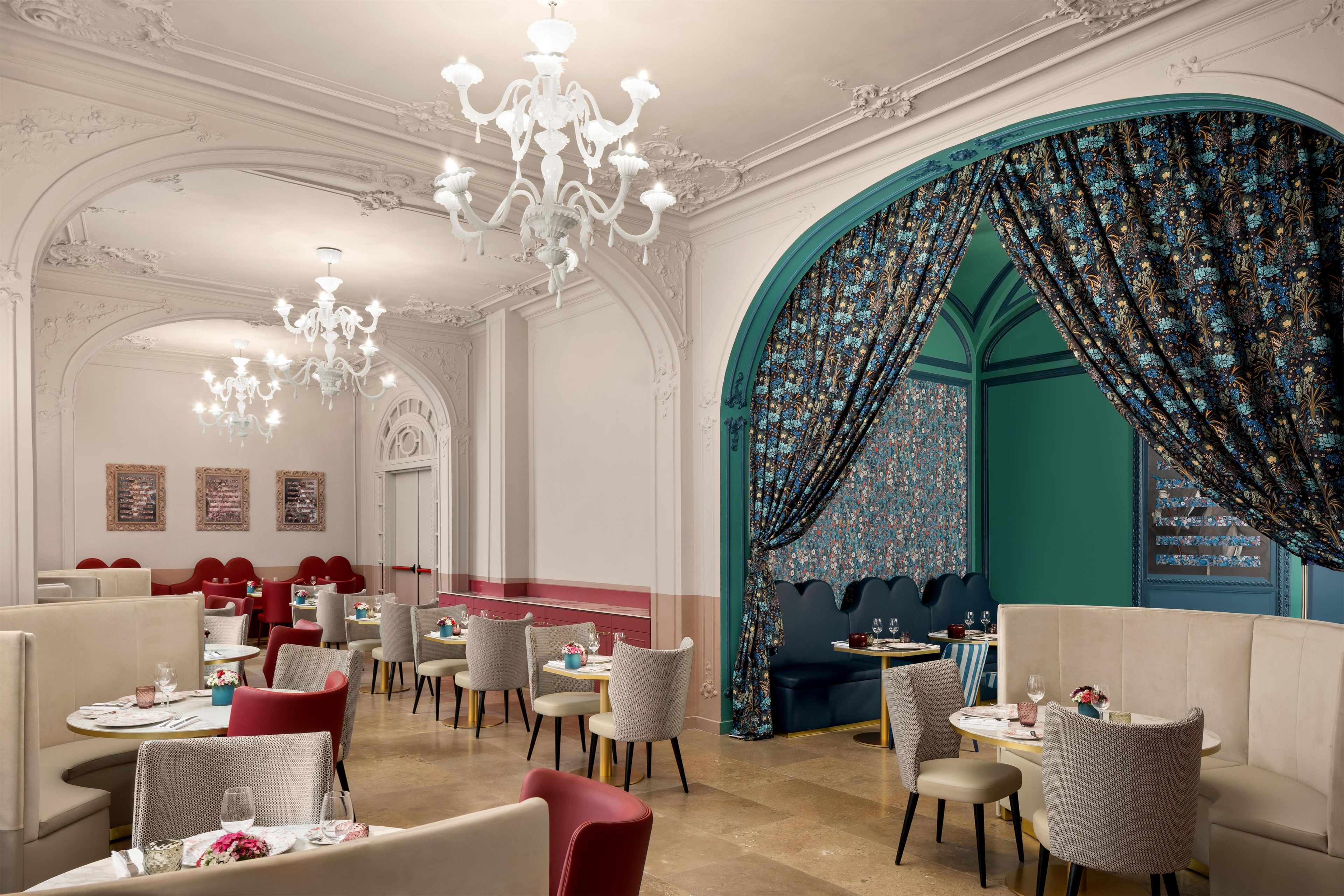 A dining area with chandeliers, red upholstered chairs, and a teal-colored wall with floral curtains.