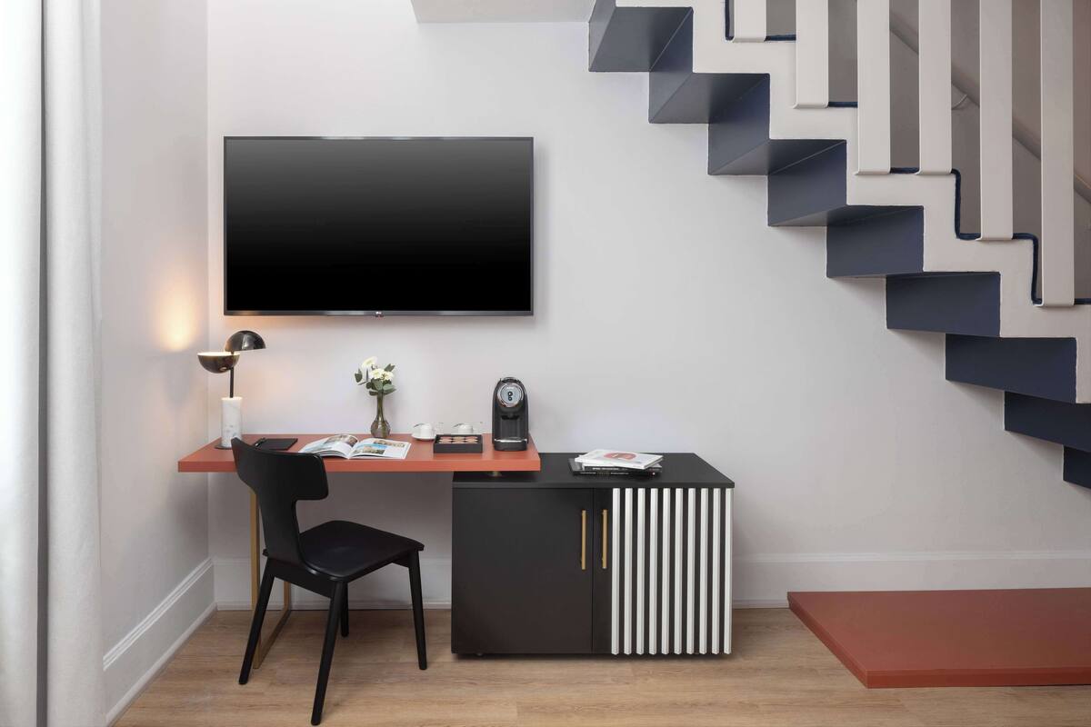 Ein modernes Zimmer mit Flachbildfernseher an der Wand, einem Schreibtisch mit Stuhl, einem kleinen Tisch mit Lampe und einer Treppe mit außergewöhnlichem Design.
