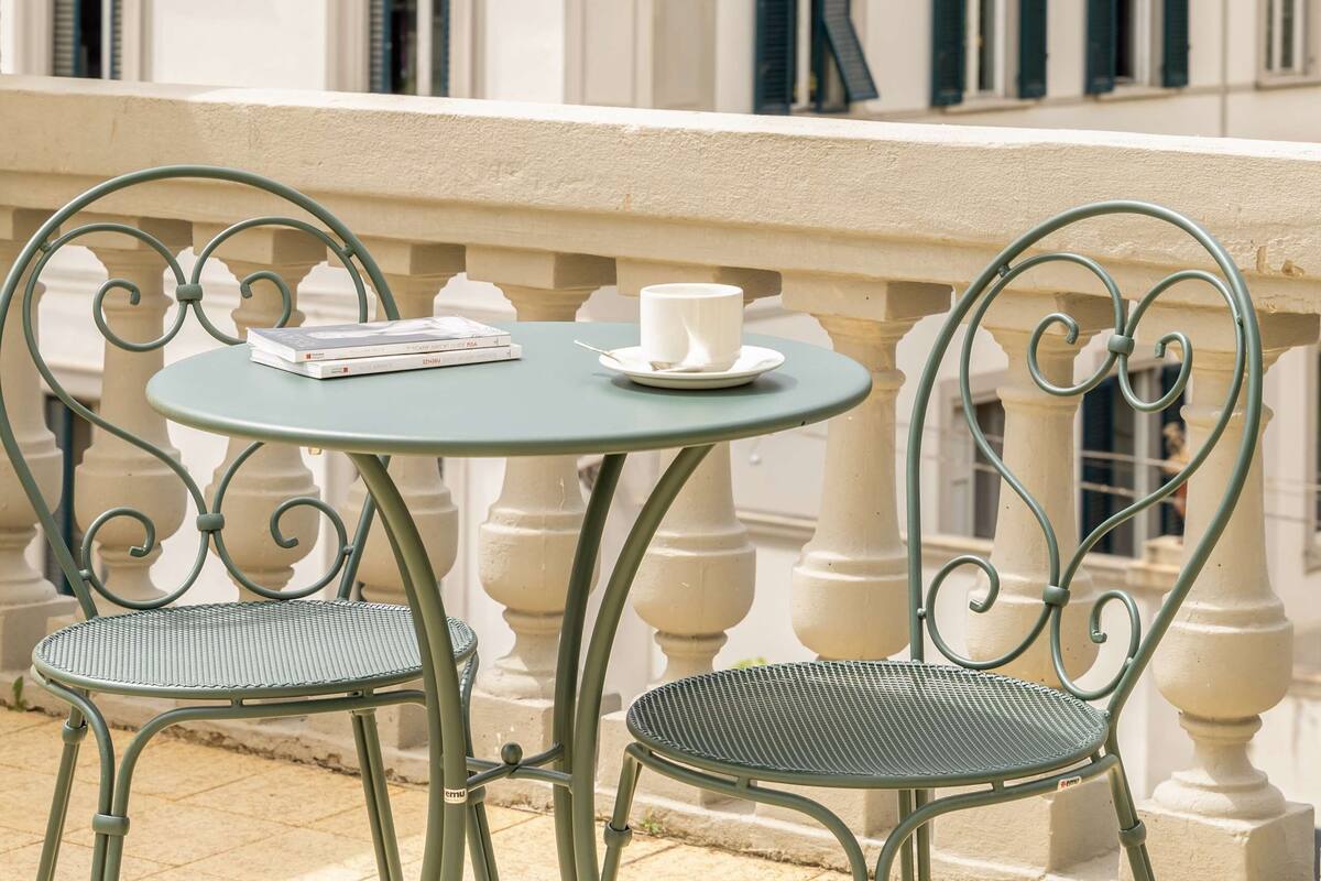 Ein Balkon mit rundem Tisch und zwei Stühlen, mit Blick auf ein Gebäude mit zahlreichen Fenstern und Fensterläden.