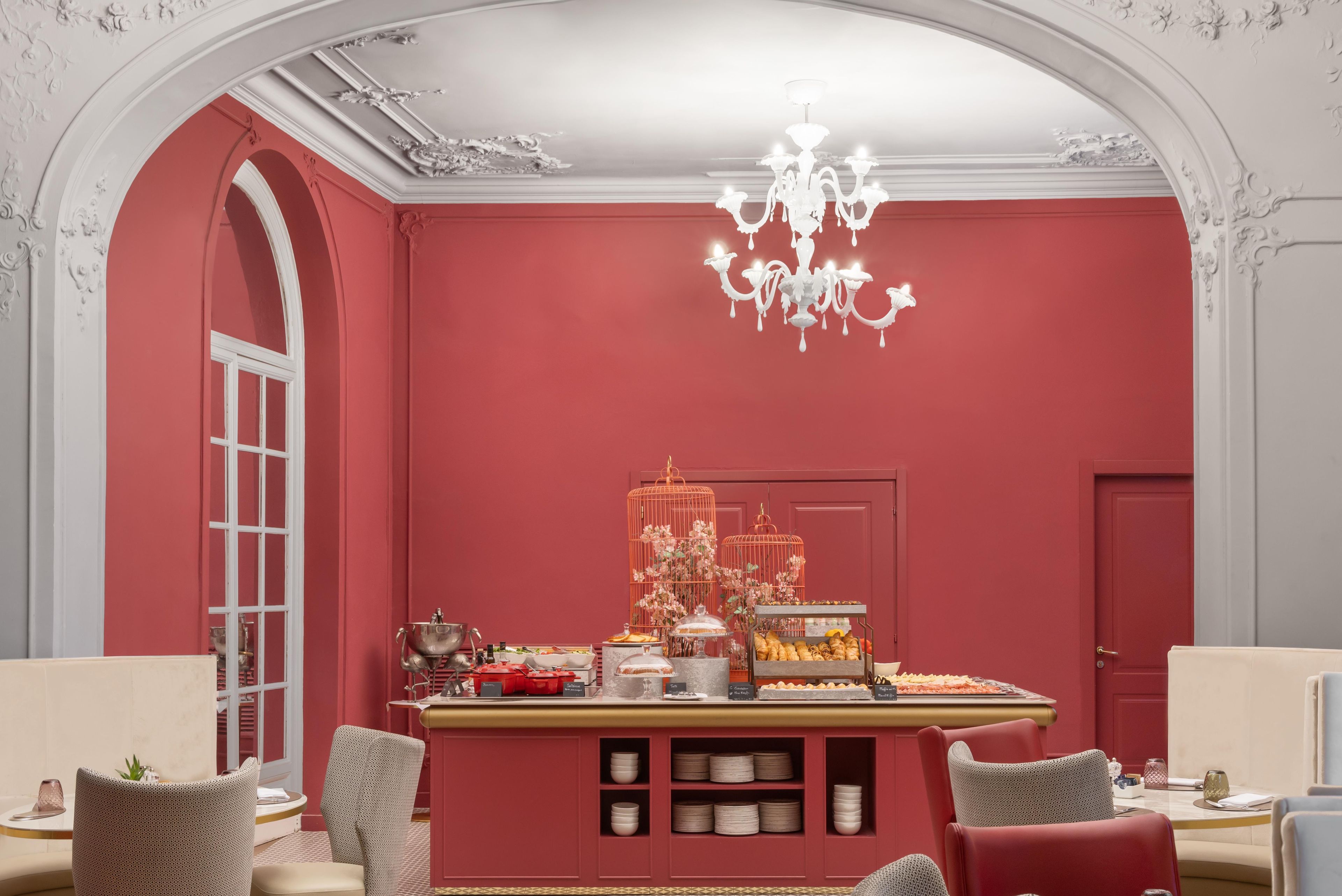 Une pièce avec un mur rouge, un lustre, une cage à oiseaux décorée de fleurs et une table de buffet.