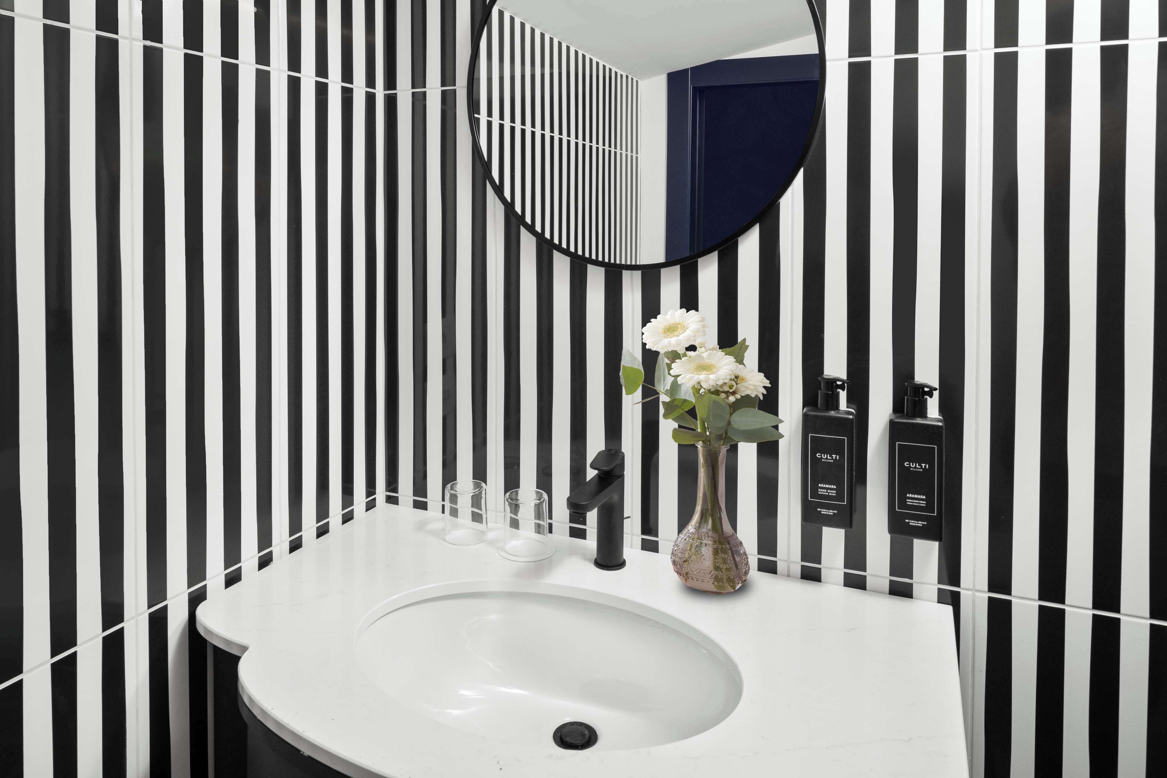 A bathroom with a white sink, black and white striped wallpaper, a round mirror, and a vase with flowers.